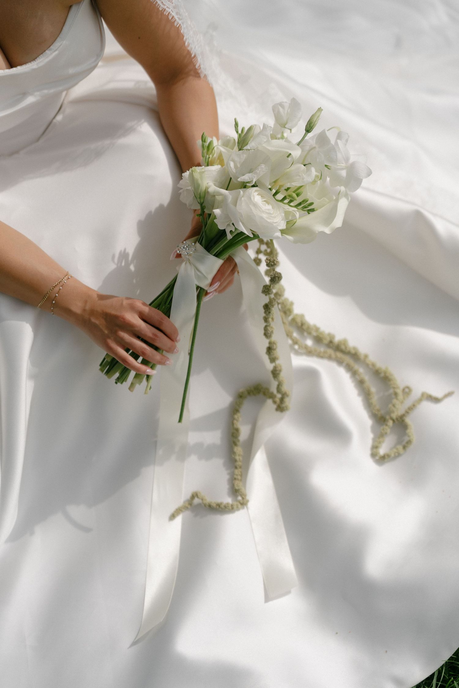 Bride holding elegant white wedding bouquet with ribbon and pearl details on white satin dress.