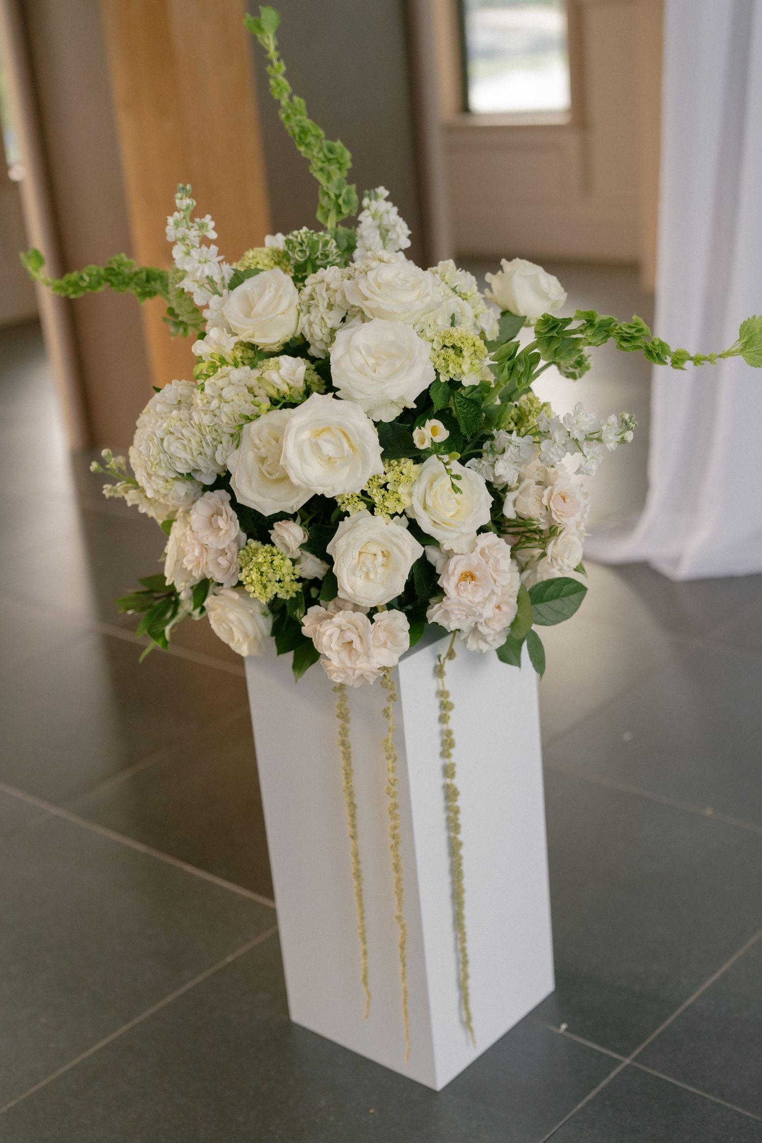 Elegant white floral arrangement with roses and greenery on a white pedestal at a wedding venue.