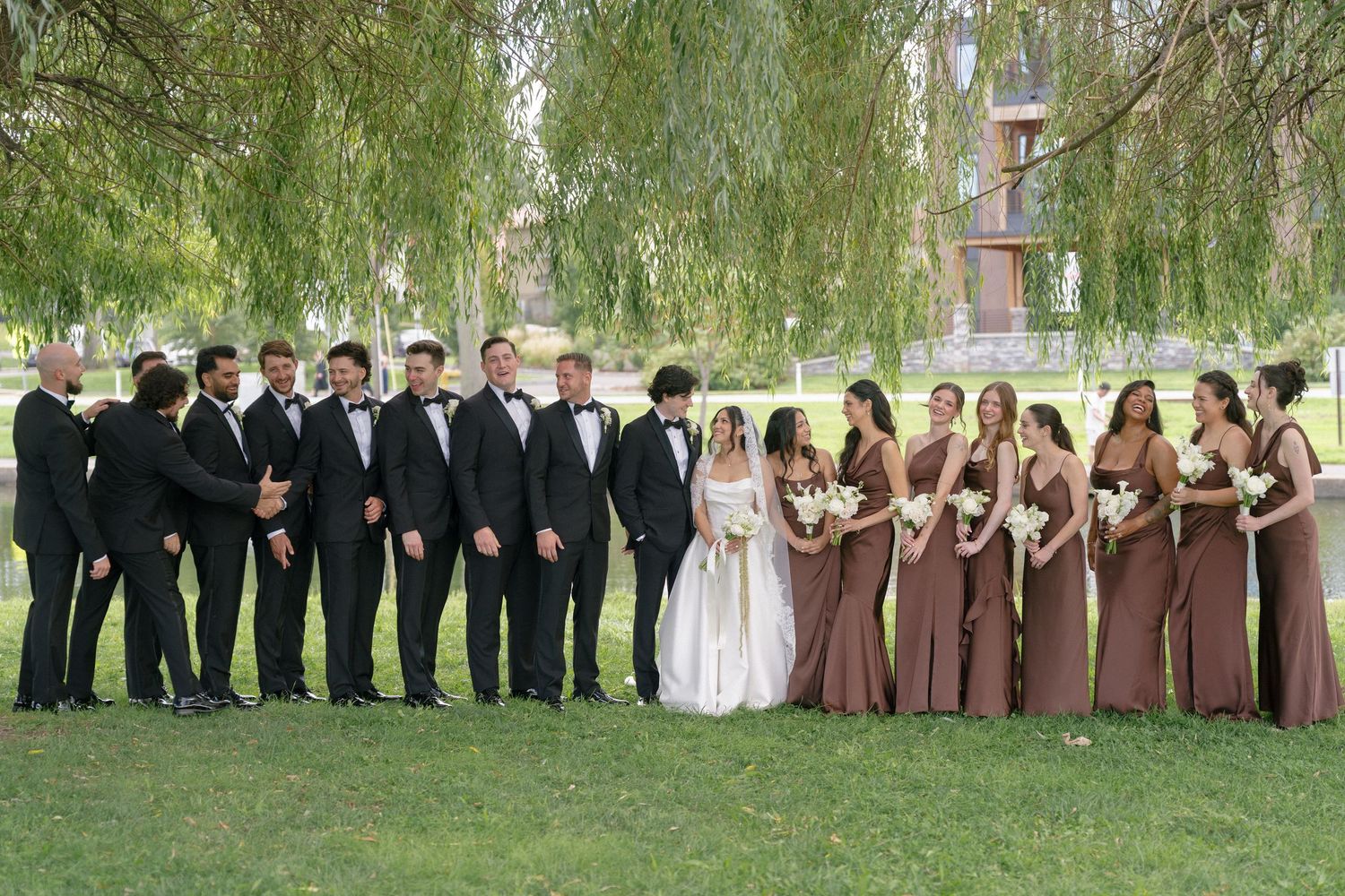Large wedding party poses outdoors with groomsmen in tuxedos and bridesmaids in brown gowns.