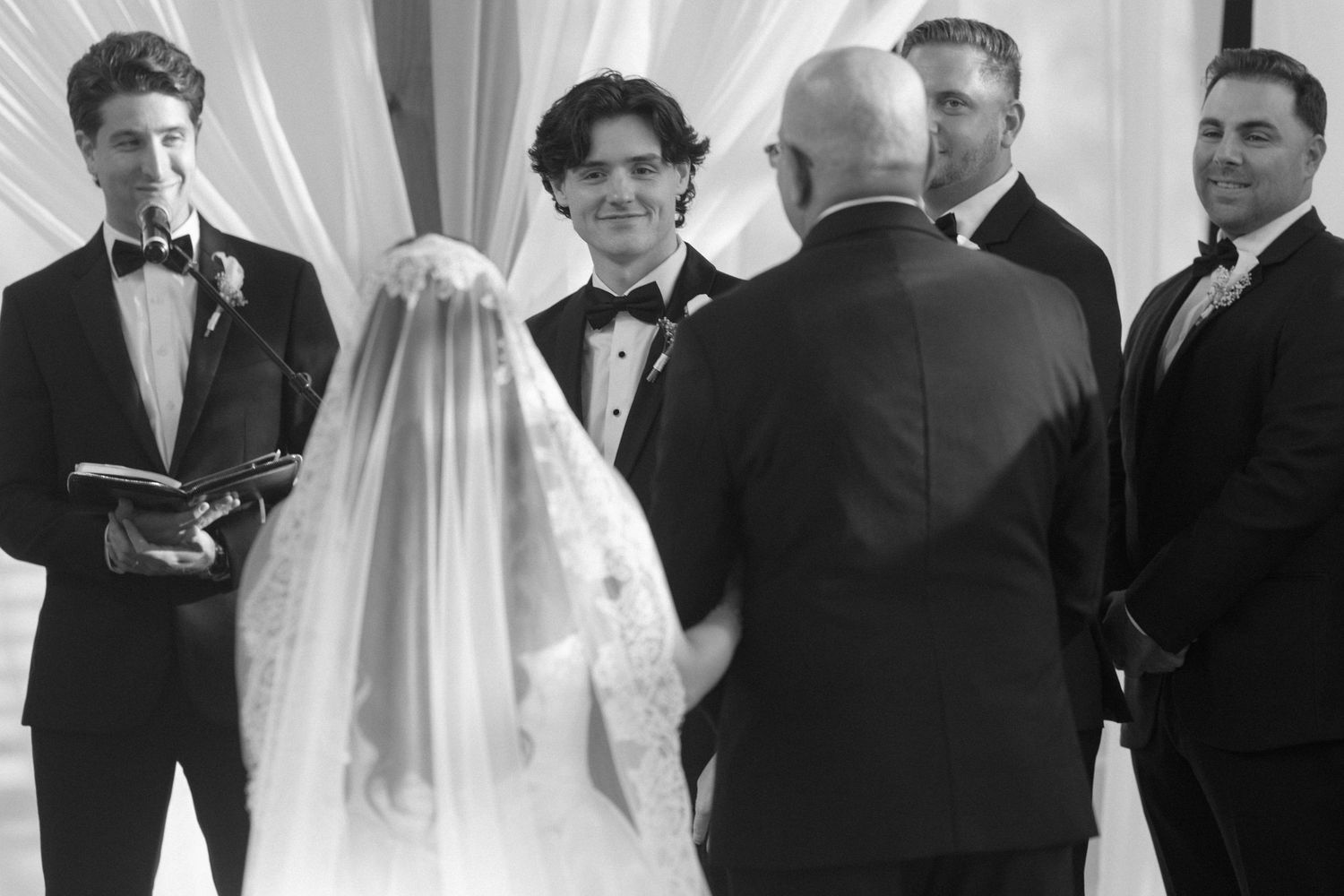 Black and white wedding ceremony photo showing bride, groom, and officiant at the altar with groomsmen standing nearby.