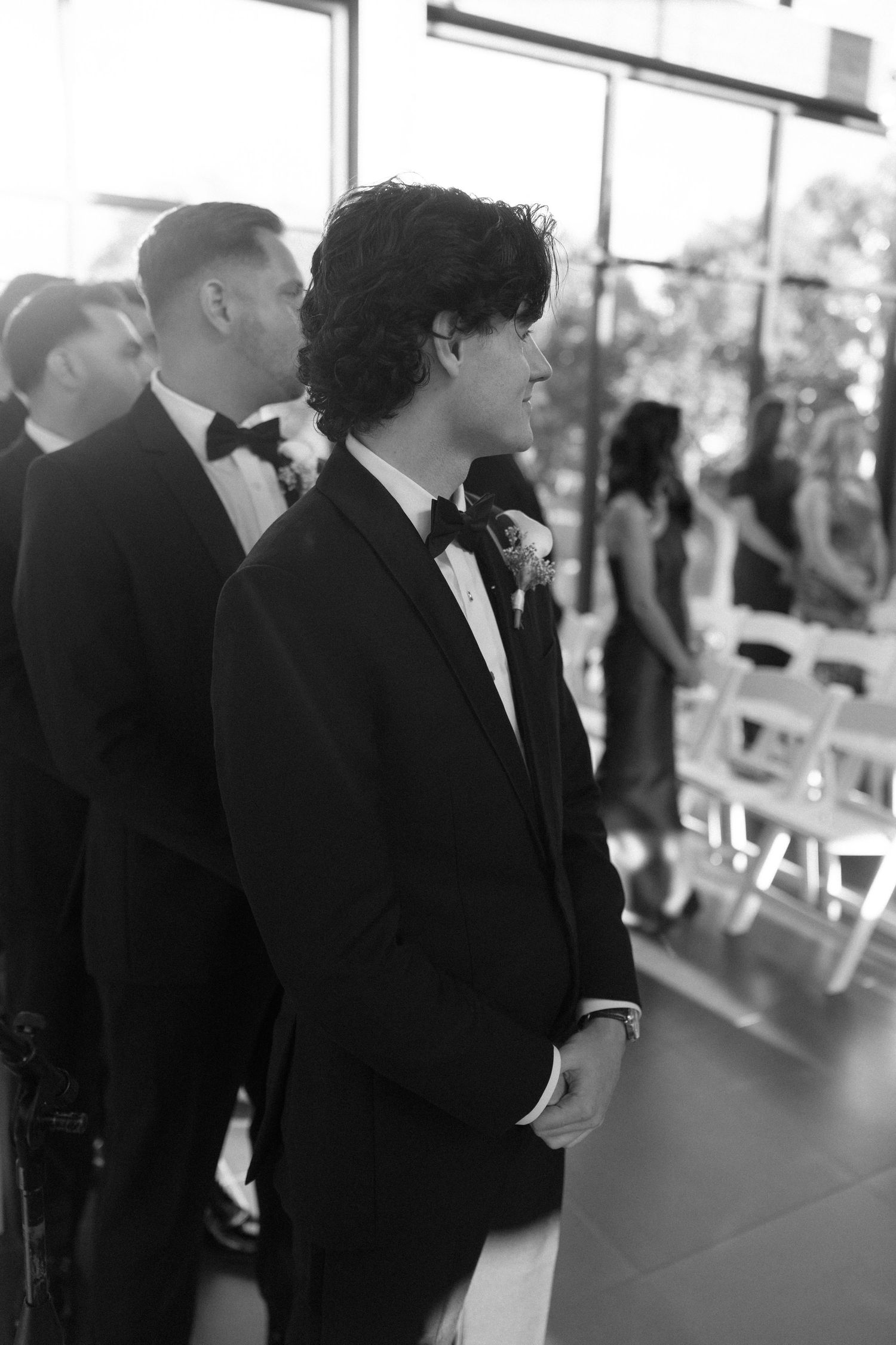 Black and white photo of a groom in a tuxedo with bow tie standing at the altar during a wedding ceremony.