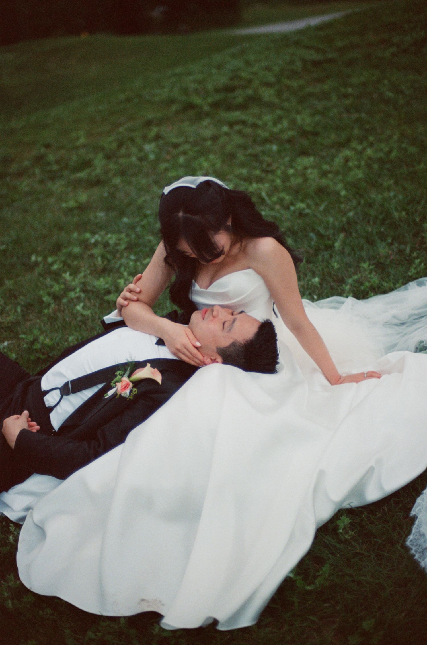 Romantic bride in white gown tenderly holds groom lying on lush green grass in outdoor portrait.