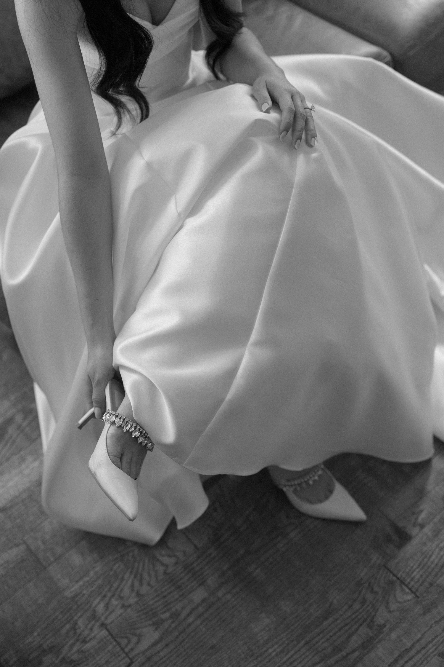 Black and white photo of bride in satin ball gown fastening crystal heeled wedding shoes while seated.