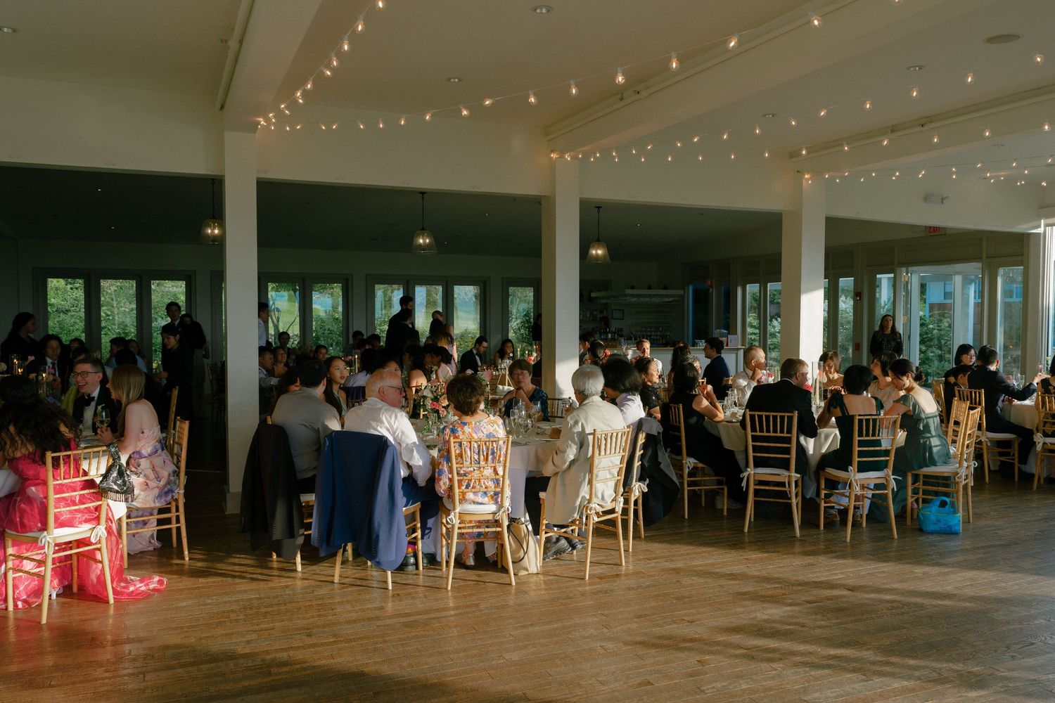 Wedding reception hall filled with guests seated at decorated tables under warm string lights.
