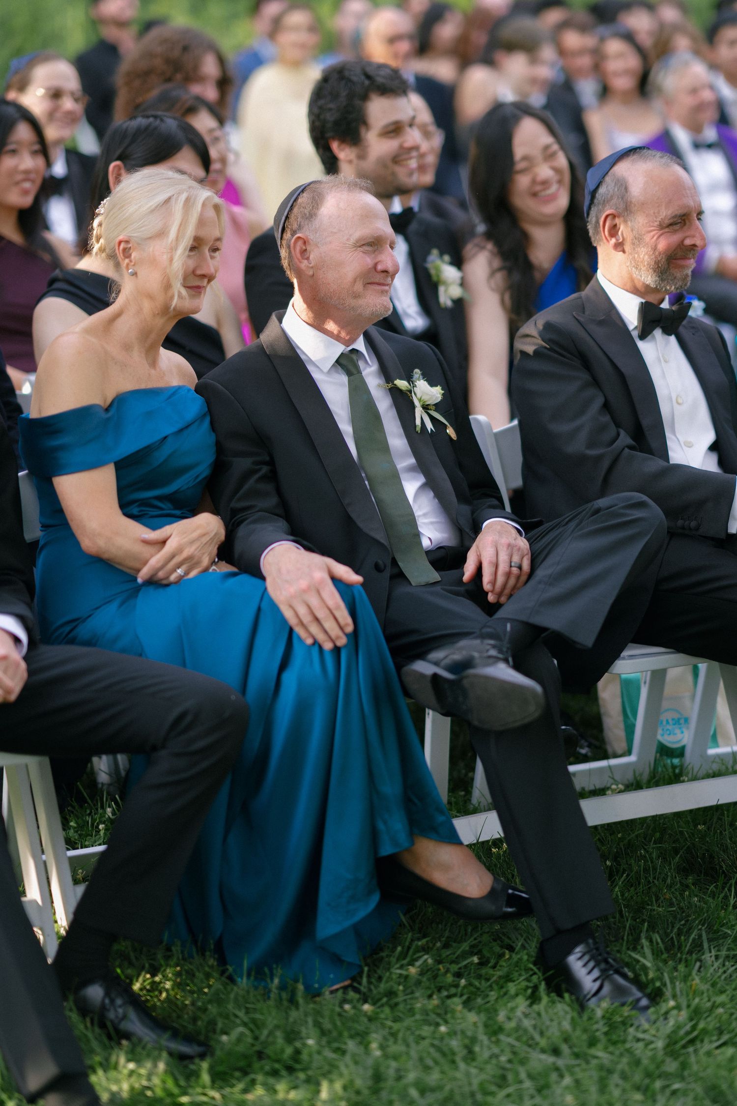 Smiling wedding guests in formal attire seated outdoors, woman in blue off-shoulder gown beside man with boutonniere.