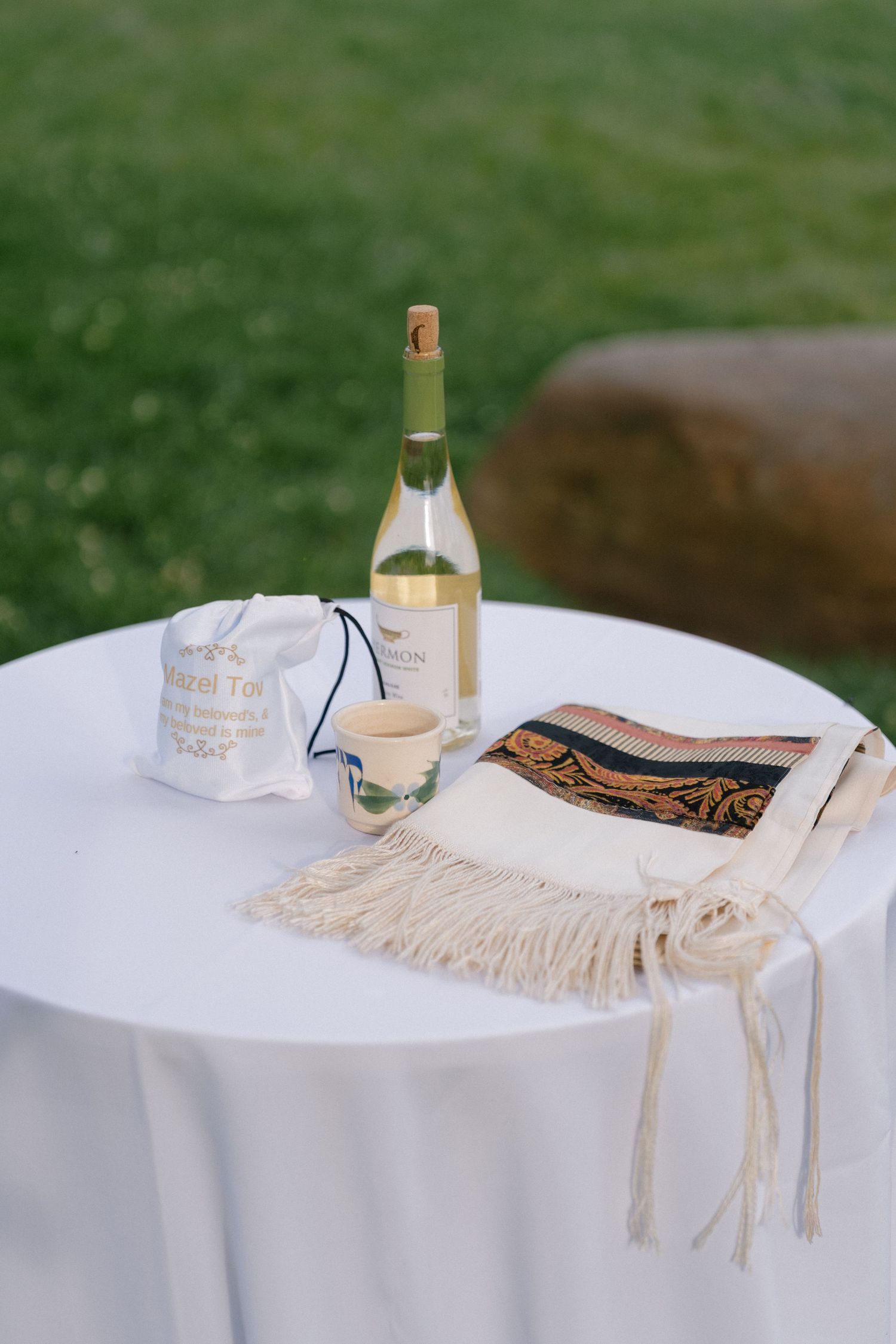 Jewish wedding table with tallit, kiddush cup, wine bottle, and mazal tov bag on white cloth outdoors.