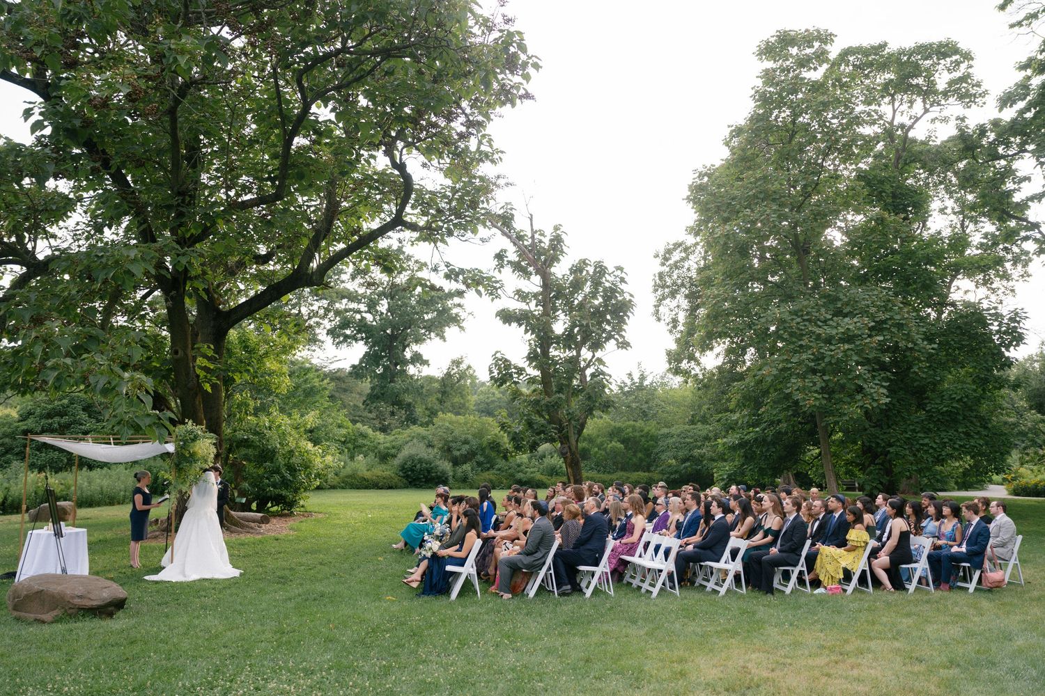 Wide-angle view of outdoor wedding ceremony with guests seated on white chairs surrounded by lush green trees.