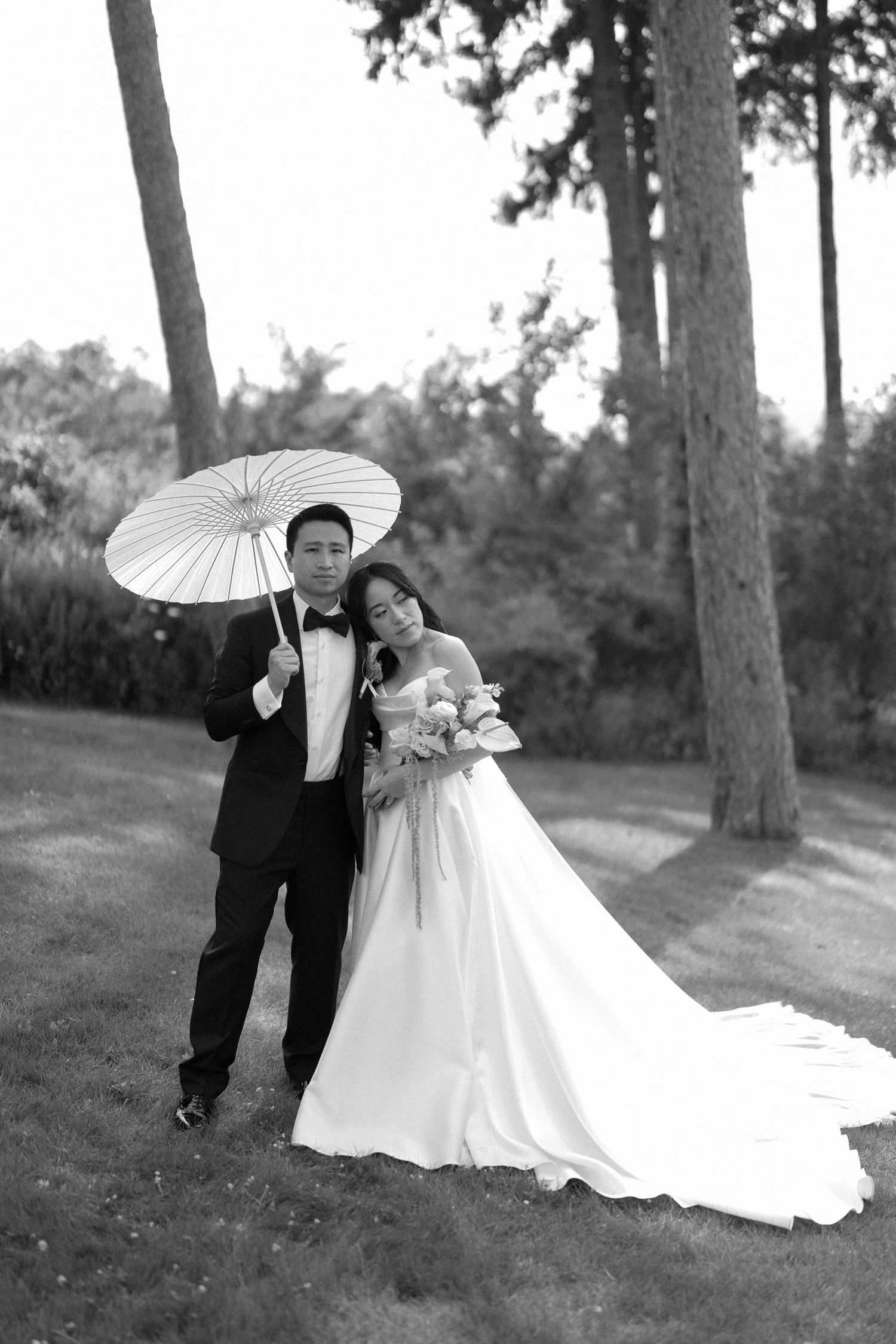 Black and white portrait of bride and groom posing with white parasol among tall pine trees.