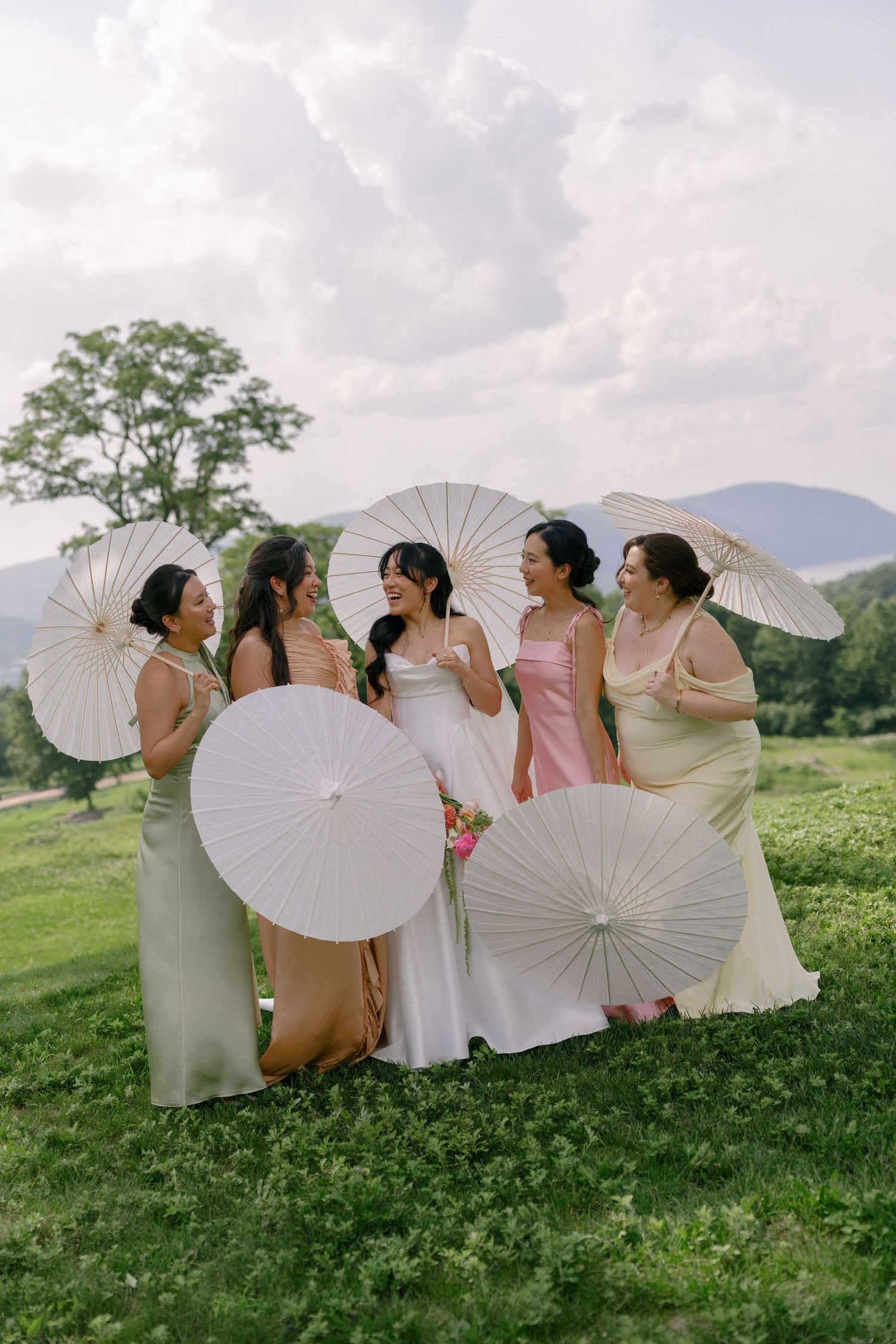 Bride and bridesmaids in colorful gowns holding white parasols on a green hillside with mountain views.
