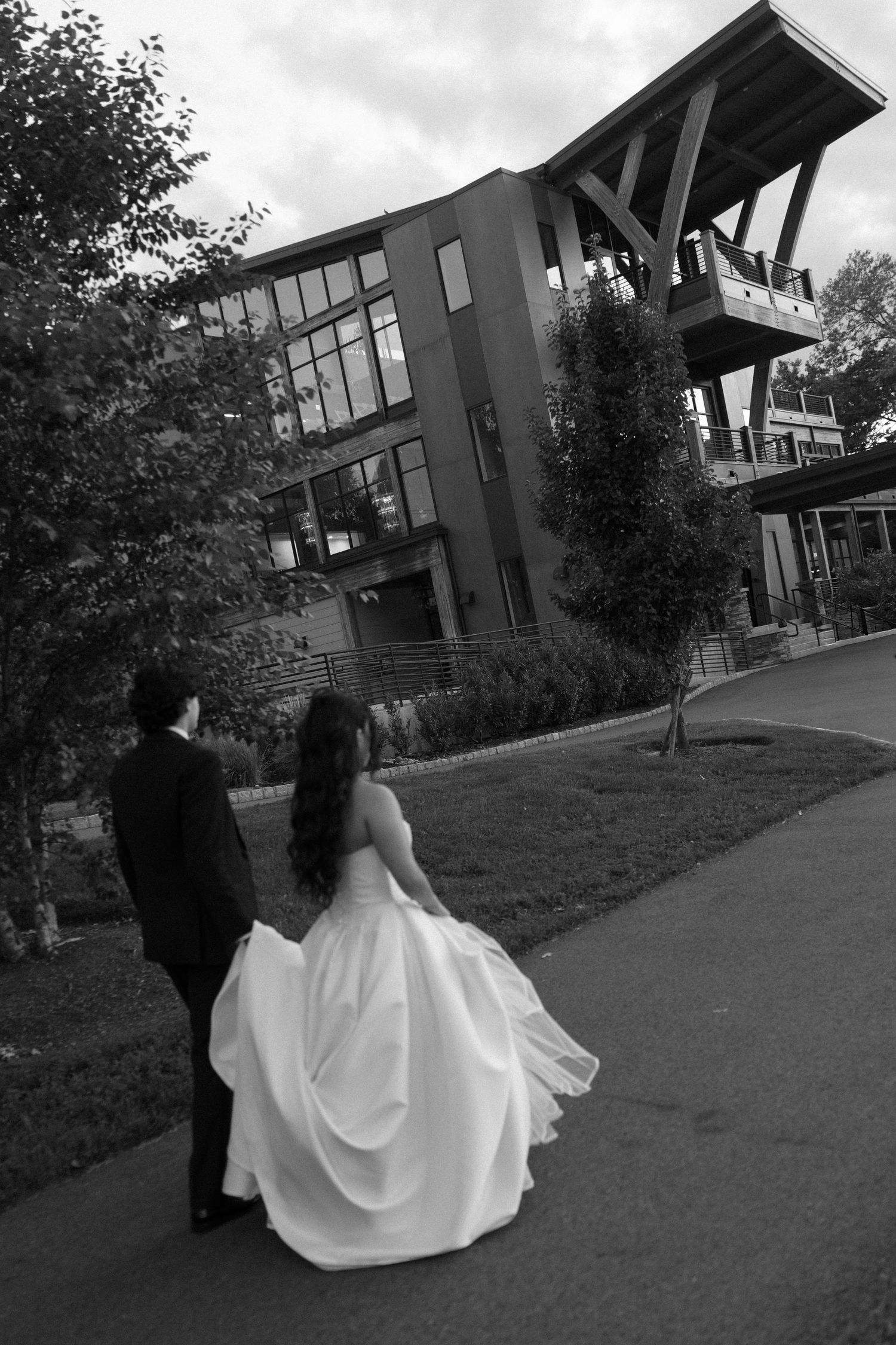 Black and white photo of bride and groom walking hand in hand toward modern wedding venue building.