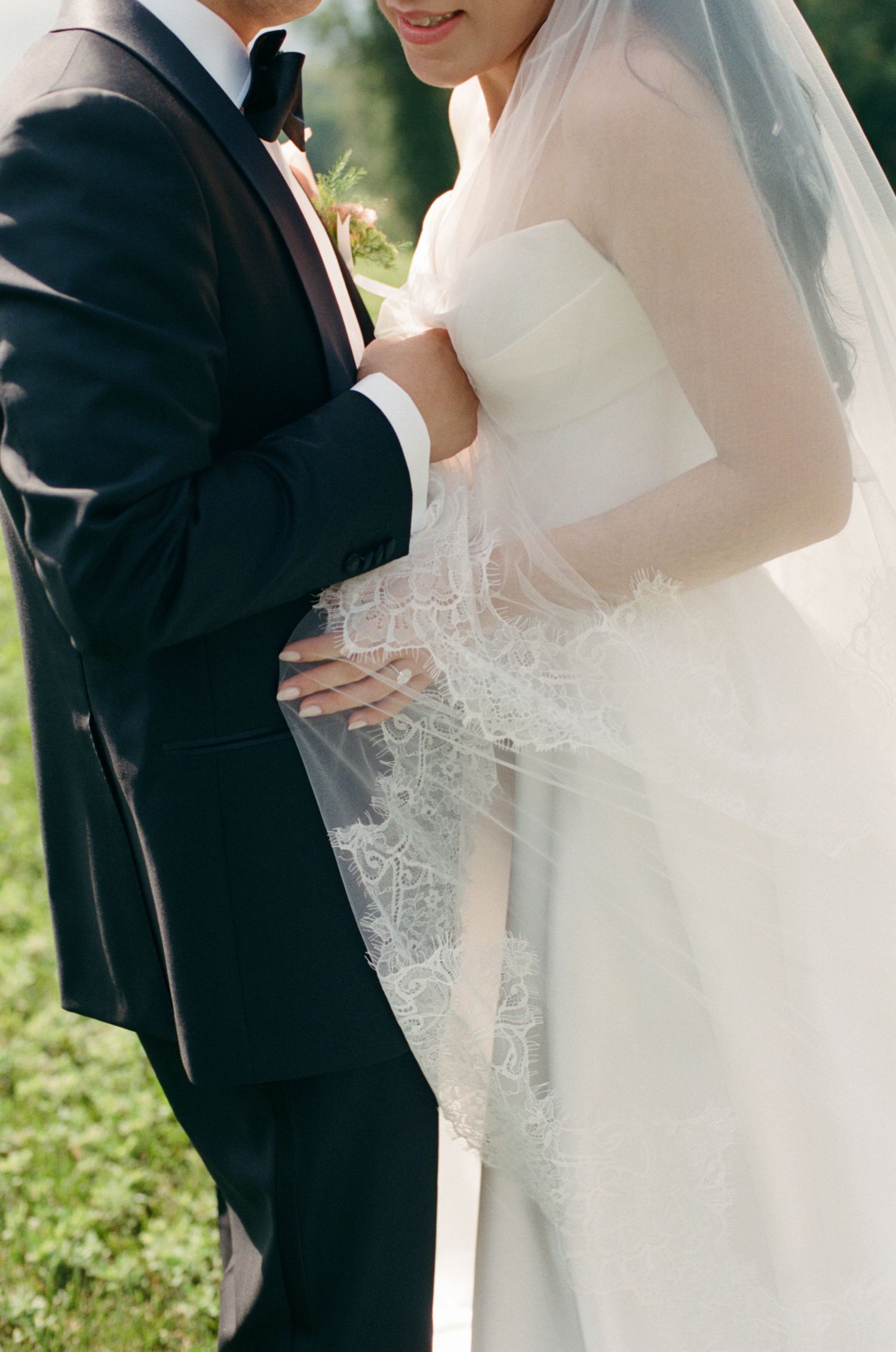 Close-up of bride in lace-trimmed veil and strapless gown embracing groom in black tuxedo outdoors.