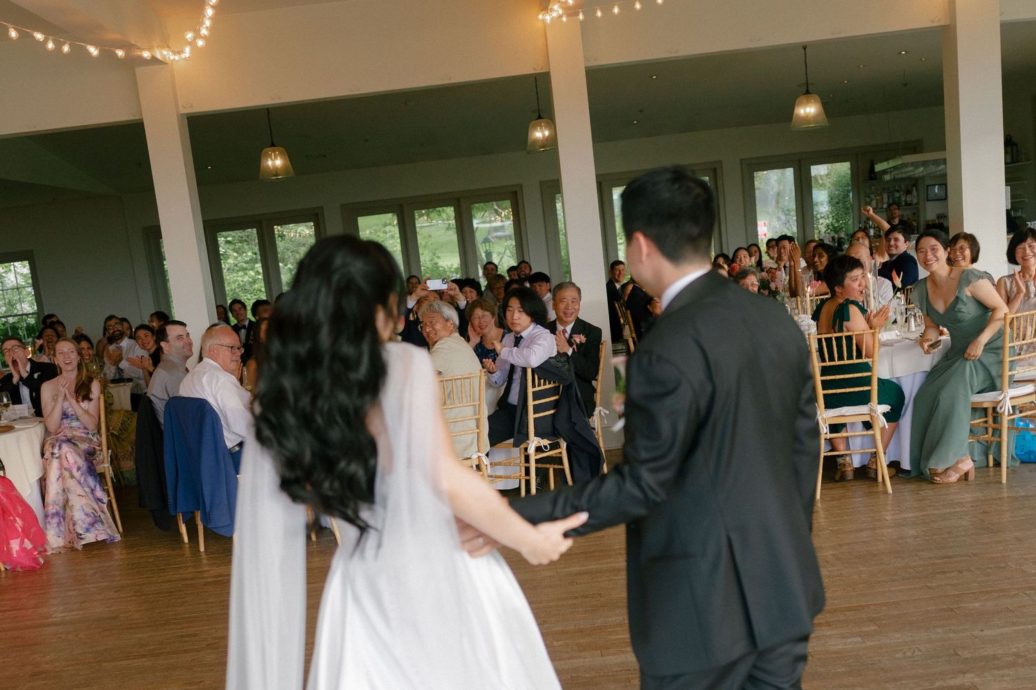Bride and groom sharing first dance before seated wedding guests in bright reception venue with large windows.