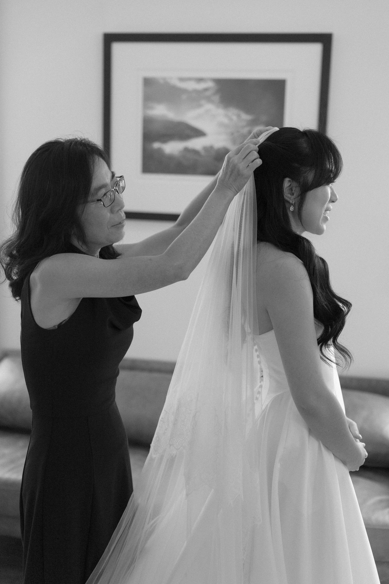 Black and white photo of woman placing veil on dark-haired bride in strapless wedding dress.