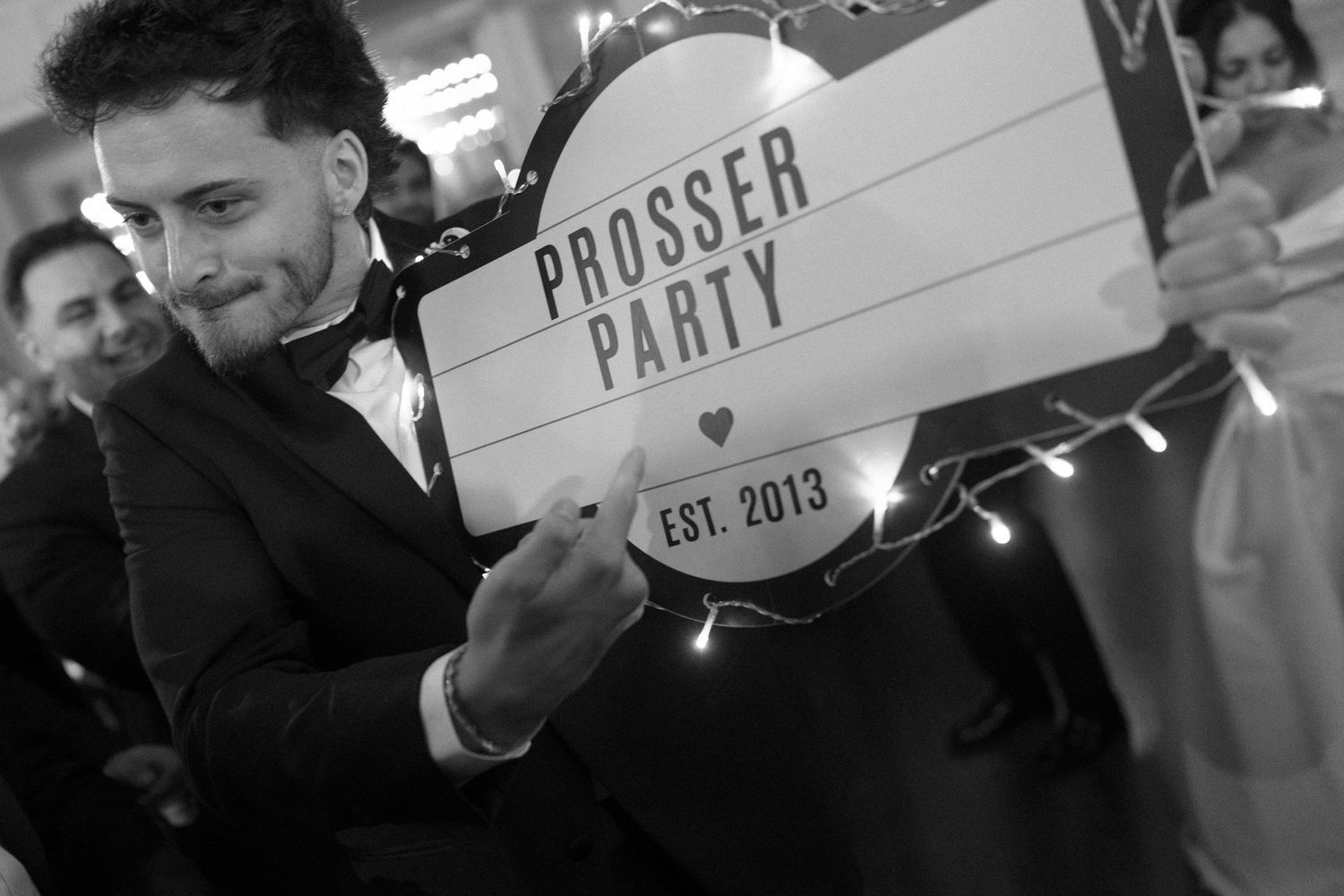 Man in tuxedo holding Prosser Party Est. 2013 light box sign at wedding reception.