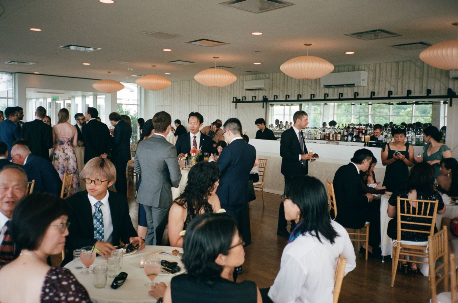 Elegant wedding reception hall filled with guests mingling at tables under globe pendant lights.