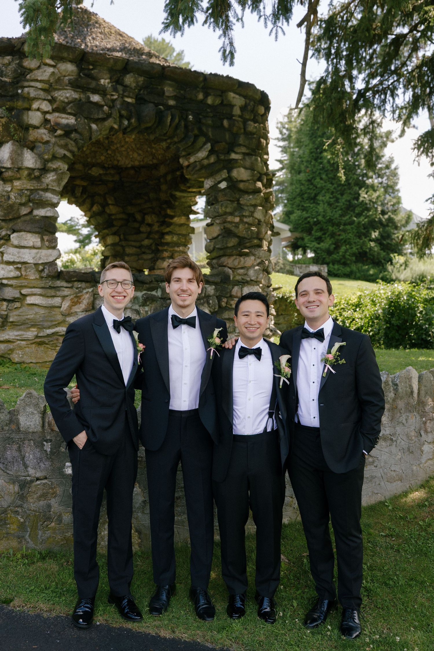Four groomsmen in black tuxedos with bow ties and boutonnieres posing outdoors near a stone wall and arch.