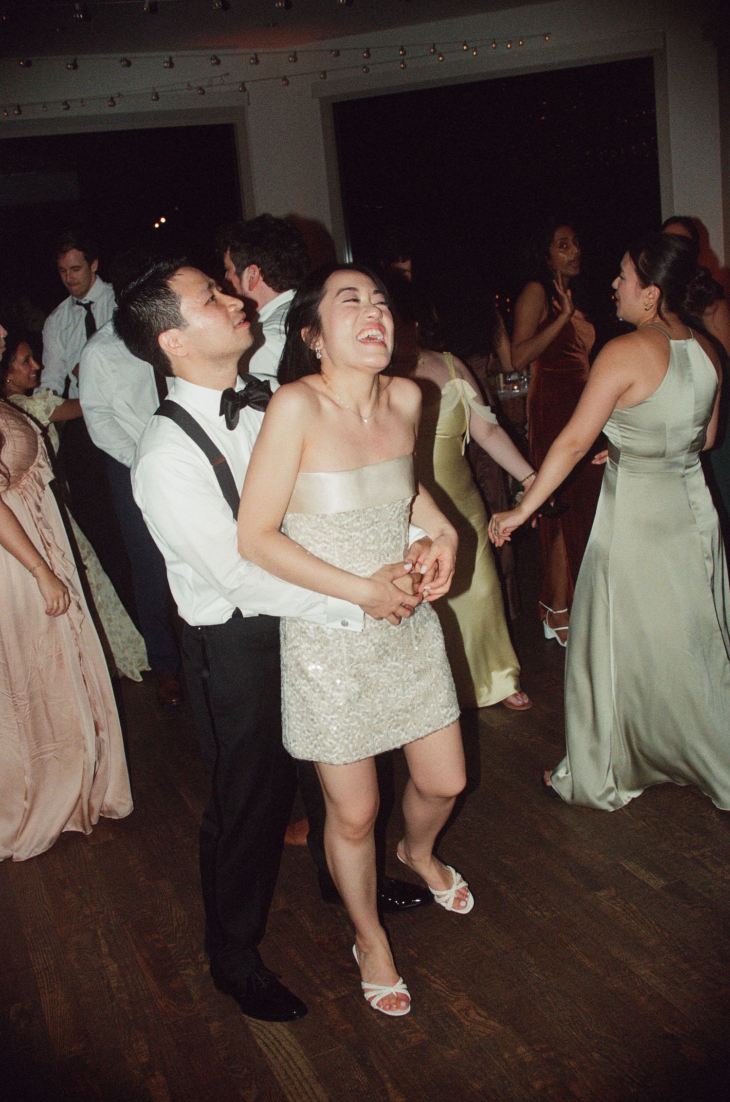 Joyful bride in sparkly mini dress laughs while dancing with groom at lively wedding reception.
