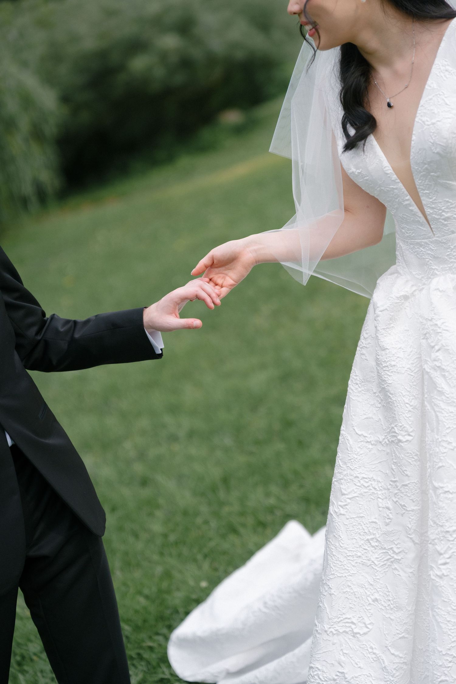 Bride and groom gently touching hands outdoors on green grass, bride wearing white lace gown and veil.