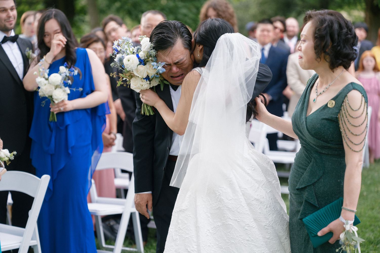 Emotional bride hugging guest during outdoor wedding ceremony, holding white floral bouquet, with family members nearby.