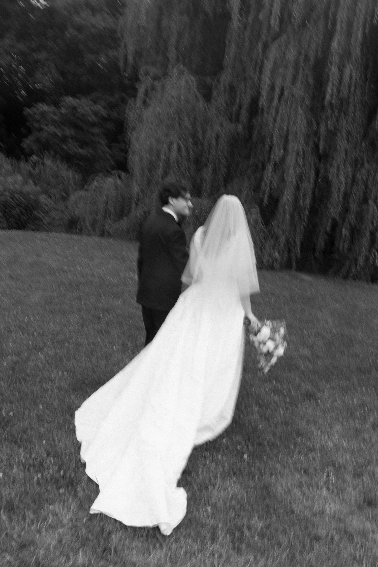 Black and white photo of bride and groom walking toward a weeping willow tree on their wedding day.