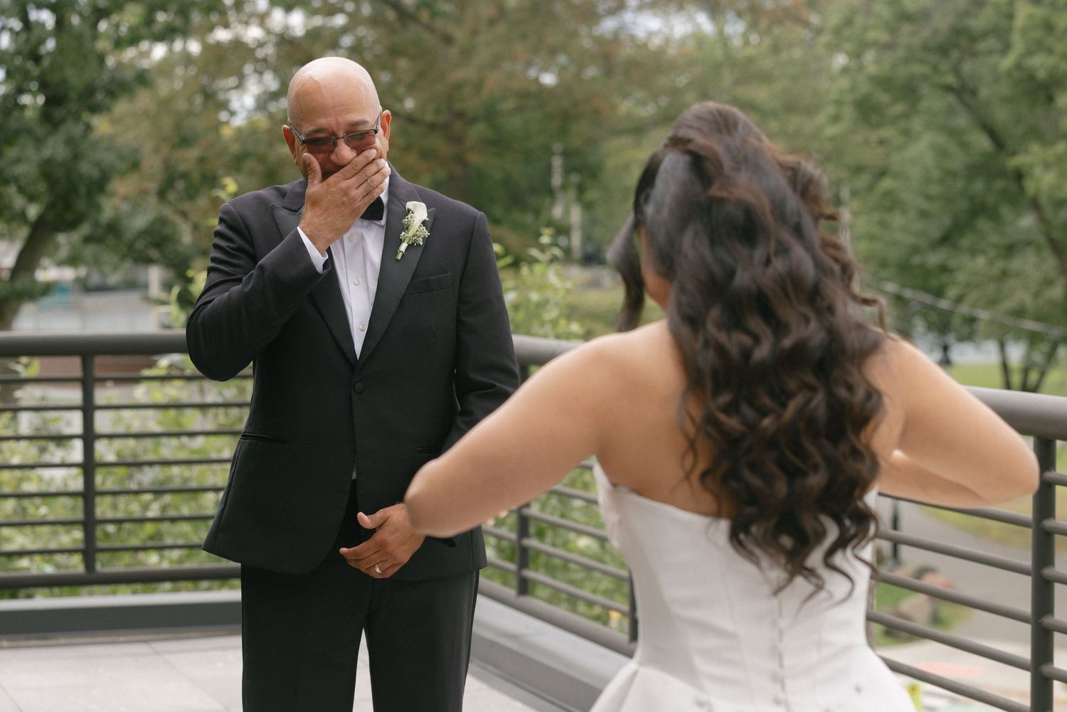 Emotional father wiping tears during first look moment with bride on bridge.