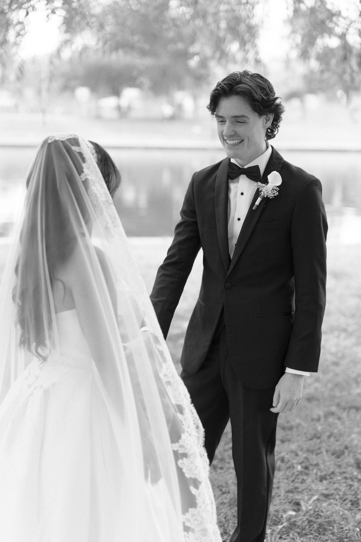 Groom smiling joyfully at bride during outdoor first look at waterside wedding venue.
