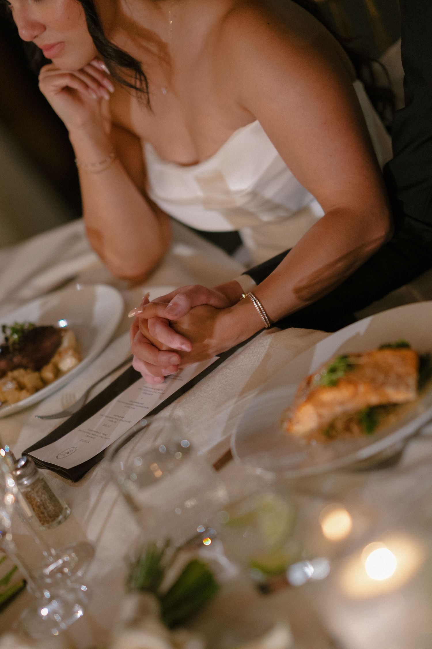 Bride at reception table with clasped hands beside dinner plate and glowing candle.