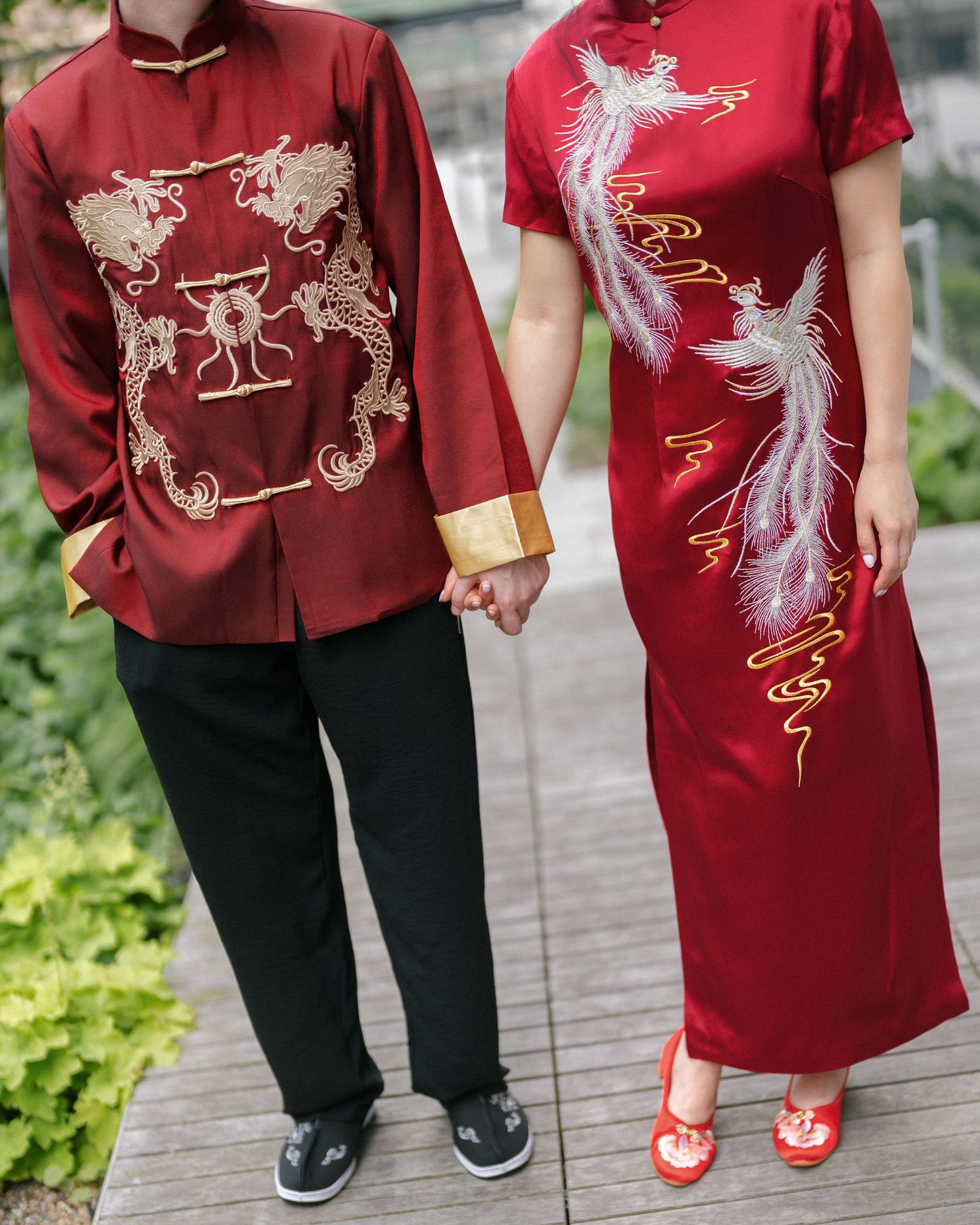 Couple in traditional red Chinese wedding attire with gold dragon and phoenix embroidery holding hands outdoors.