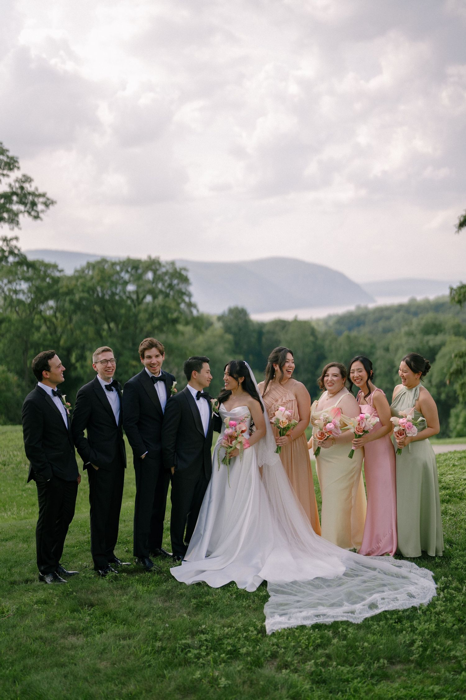 Wedding party in black tuxedos and colorful bridesmaid dresses posing on green lawn with mountain backdrop.