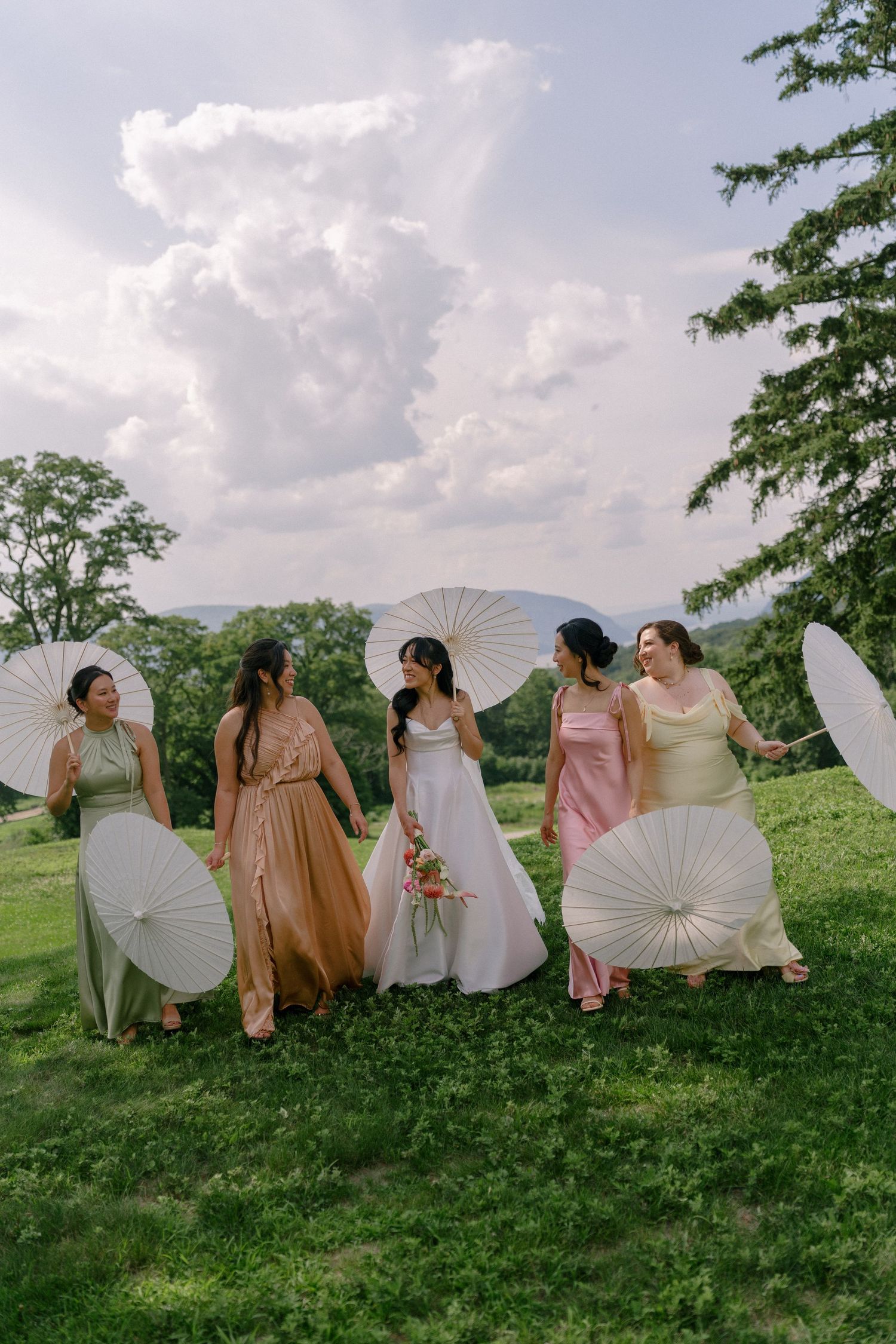 Bride and four bridesmaids in colorful mismatched dresses holding white parasols on lush green hillside.