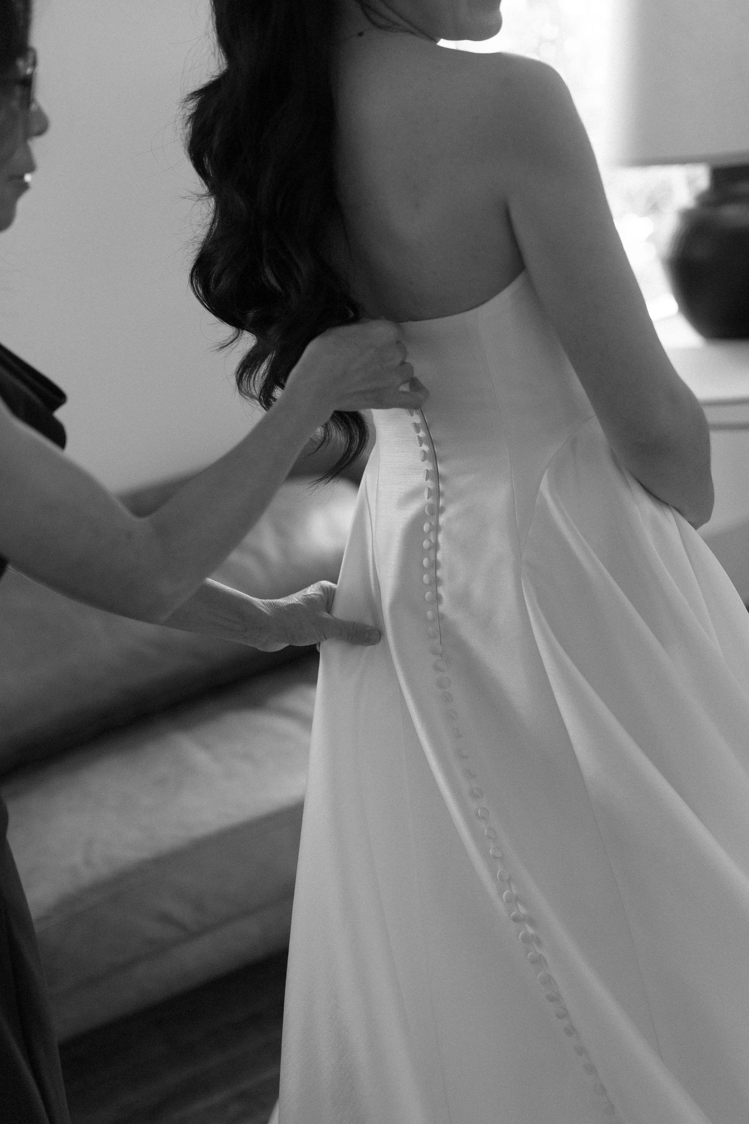 Black and white photo of bridesmaid buttoning bride's elegant strapless wedding gown with pearl buttons.