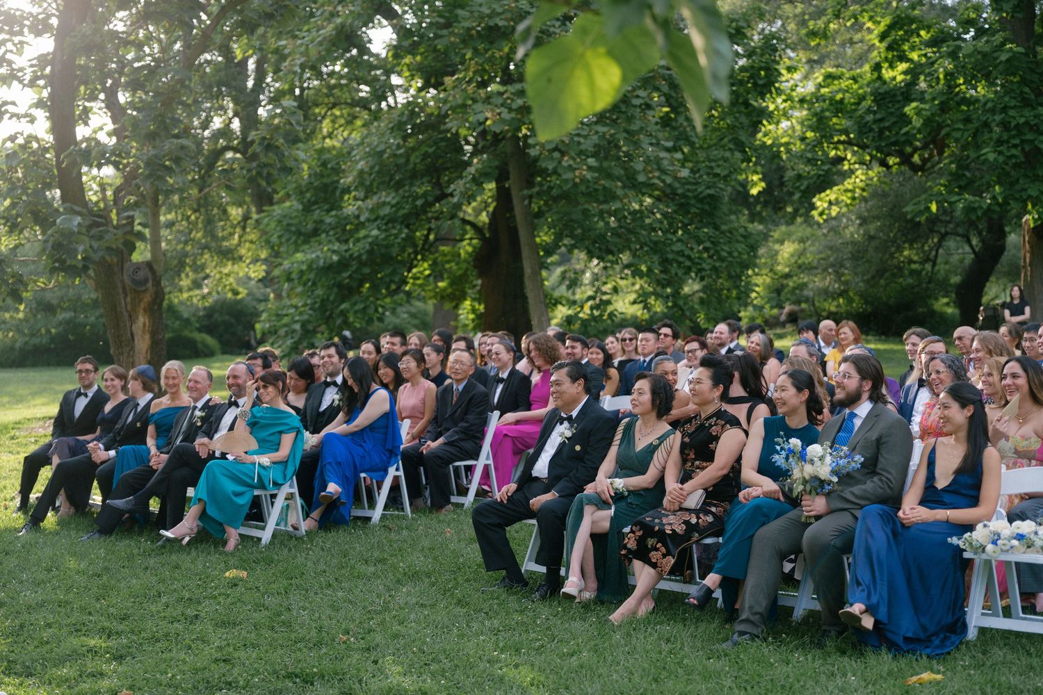 Large group of wedding guests seated outdoors in white chairs, surrounded by tall green trees during a ceremony.