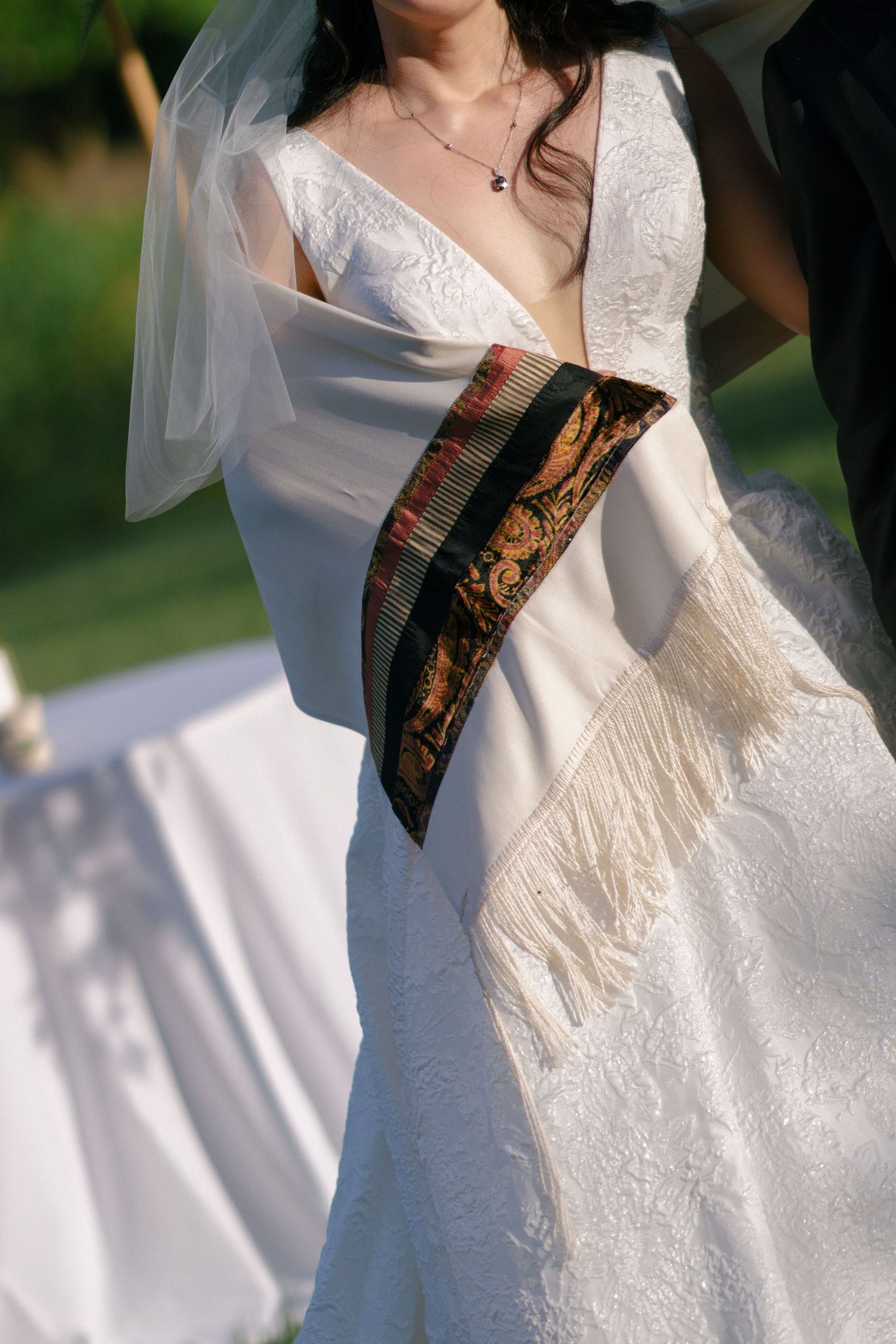 Close-up of bride in white lace wedding dress adorned with traditional decorative sash and fringe.