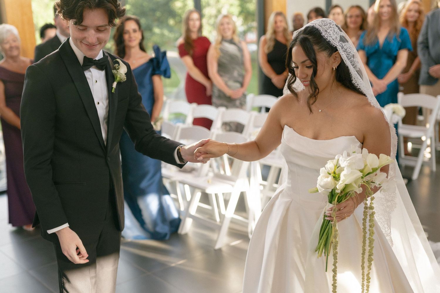 Newlywed couple walks down the aisle smiling, bride in strapless gown holding bouquet, groom in black tuxedo.