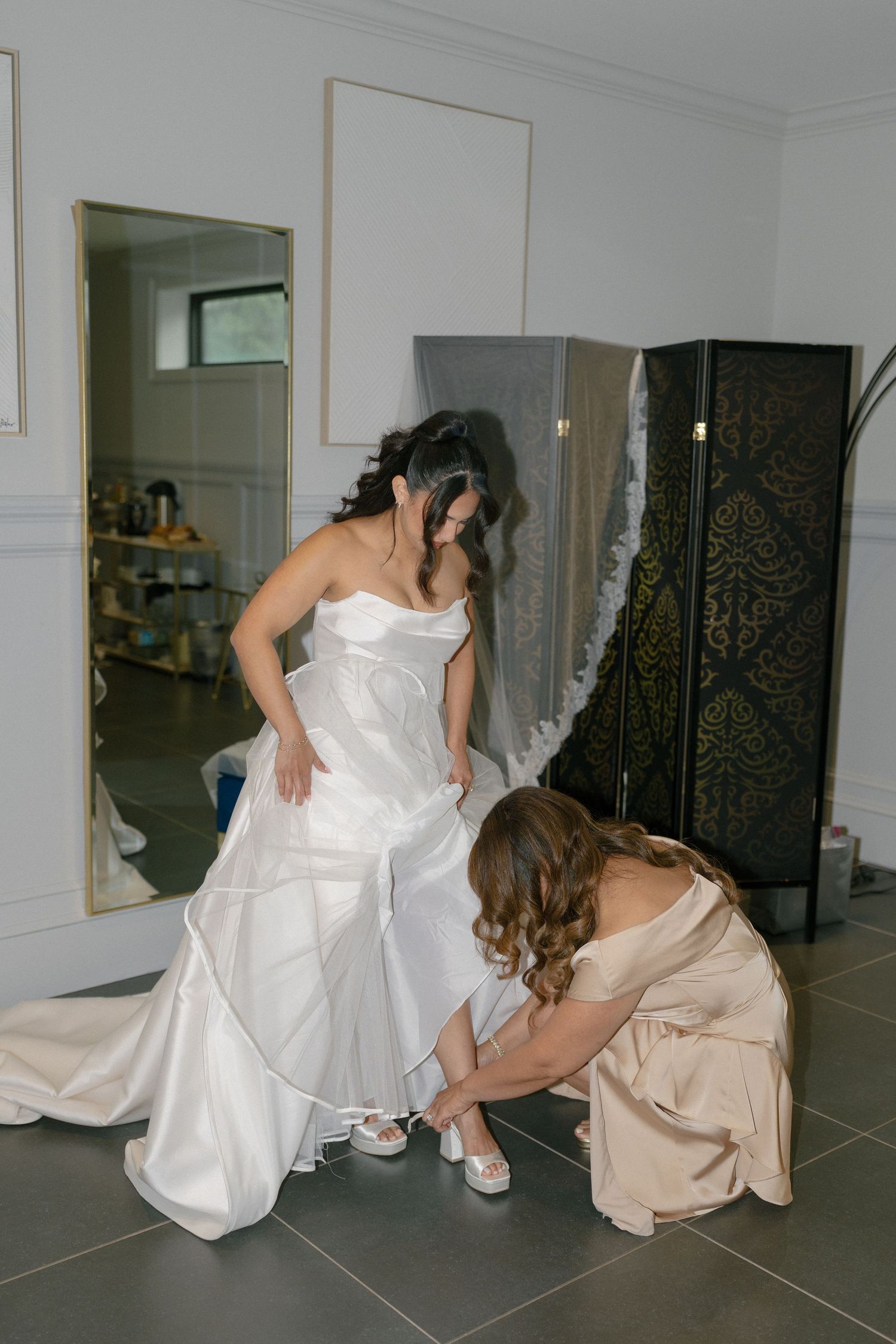Bridesmaid in champagne dress helps bride in white strapless gown with her shoes in a bright dressing room.