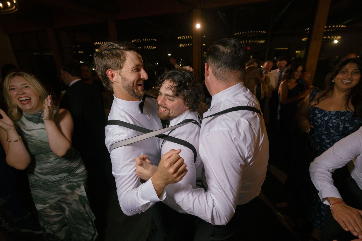 Groomsmen in white shirts and suspenders dancing joyfully at wedding reception.