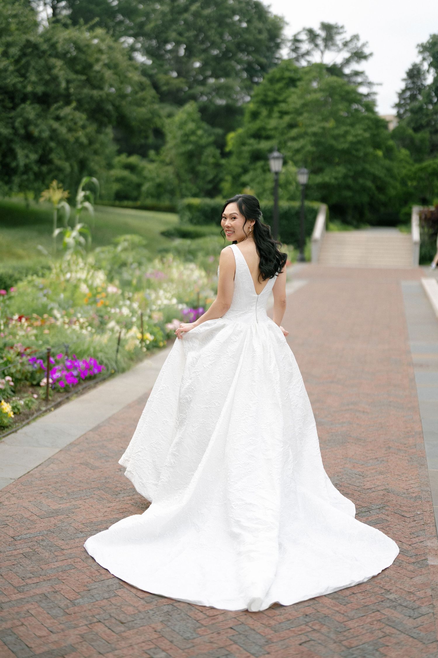 Bride in elegant white ball gown twirling on a brick path in a lush garden.