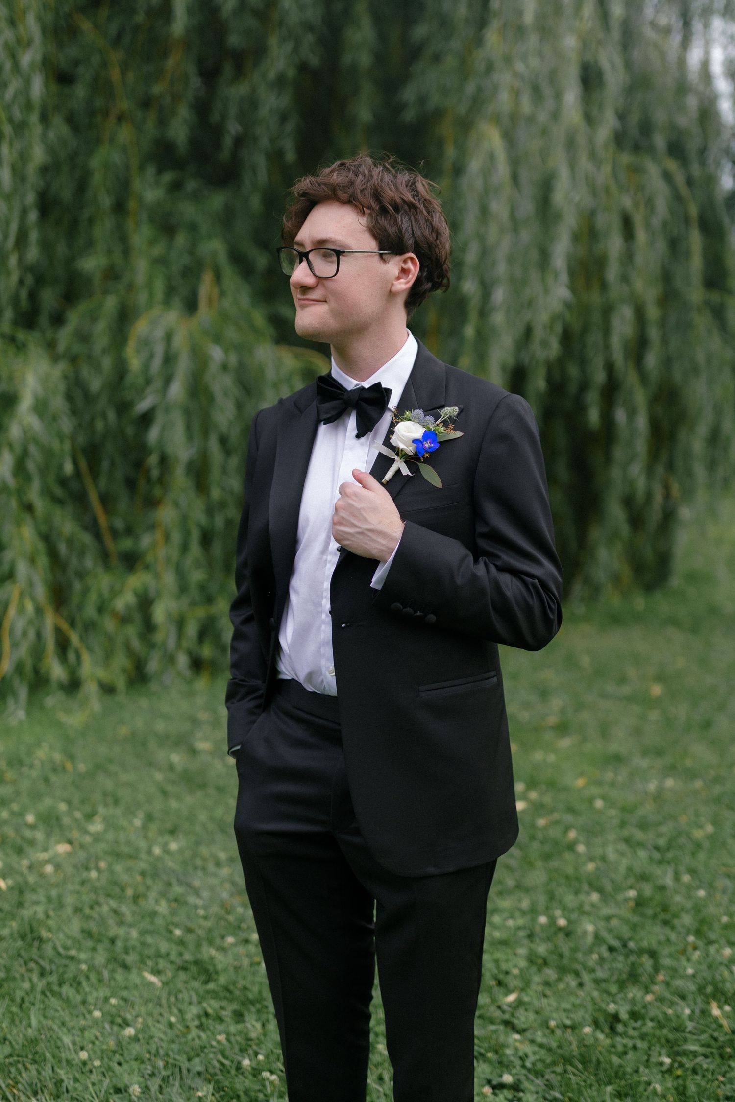 A groom in a black tuxedo with bow tie and boutonniere stands outdoors near a weeping willow tree.