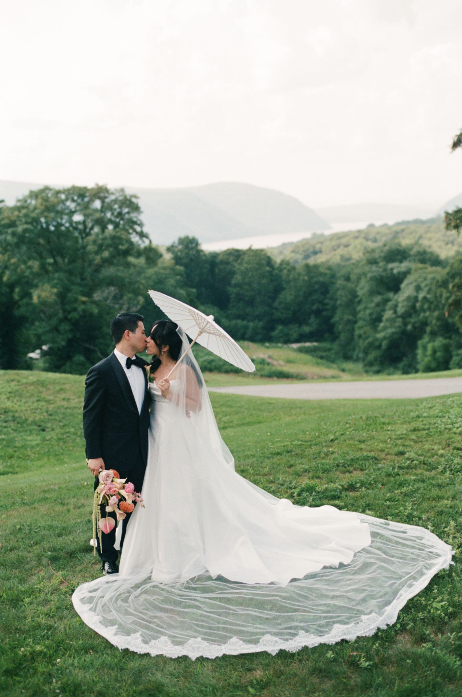 Bride and groom kissing on lush green lawn, bride holding parasol with long lace veil trailing, mountains in background.