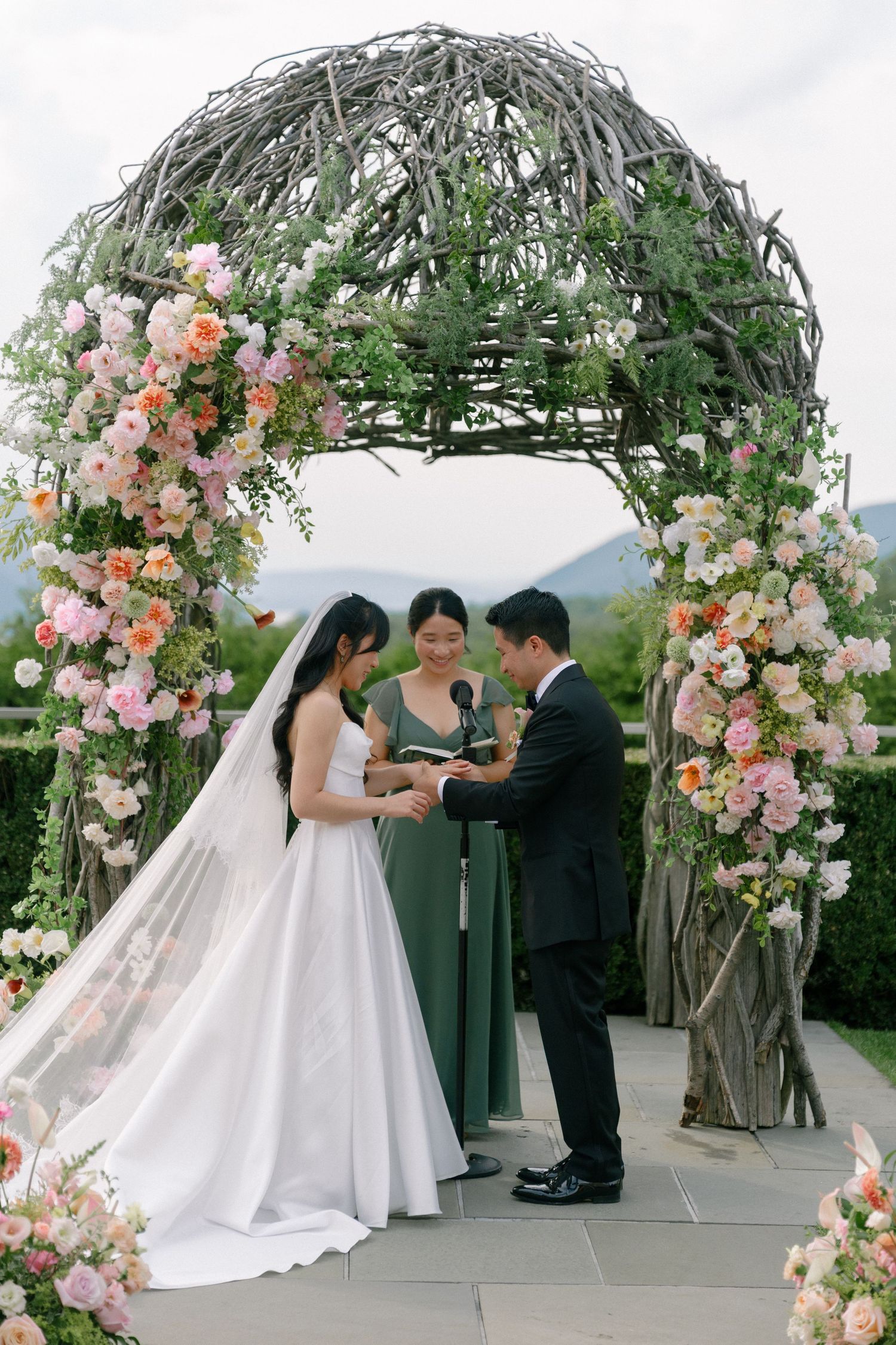 Couple exchanges rings under a floral arch adorned with pink roses at outdoor ceremony.