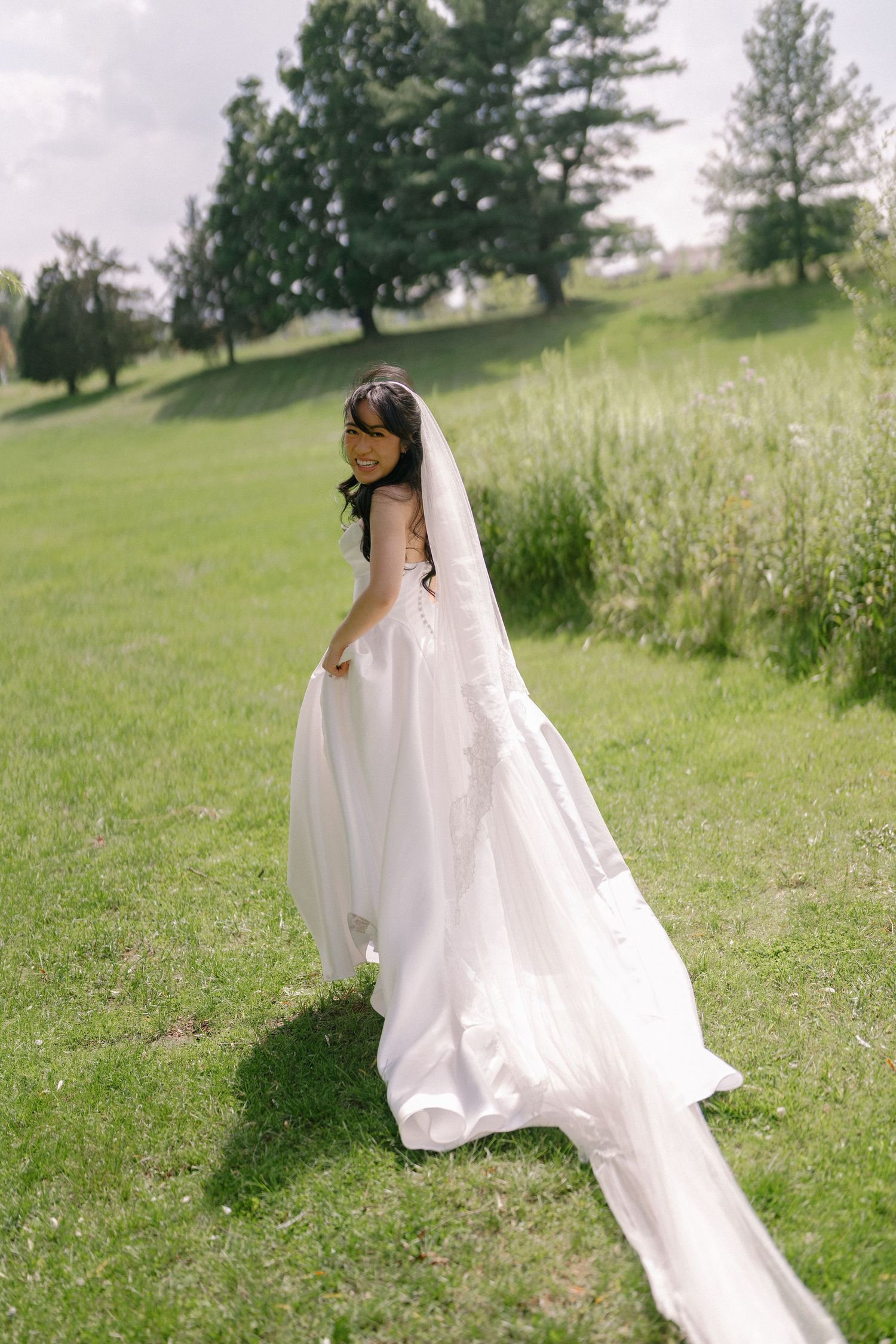 Bride in flowing white gown with long veil walks through lush green meadow on her wedding day.
