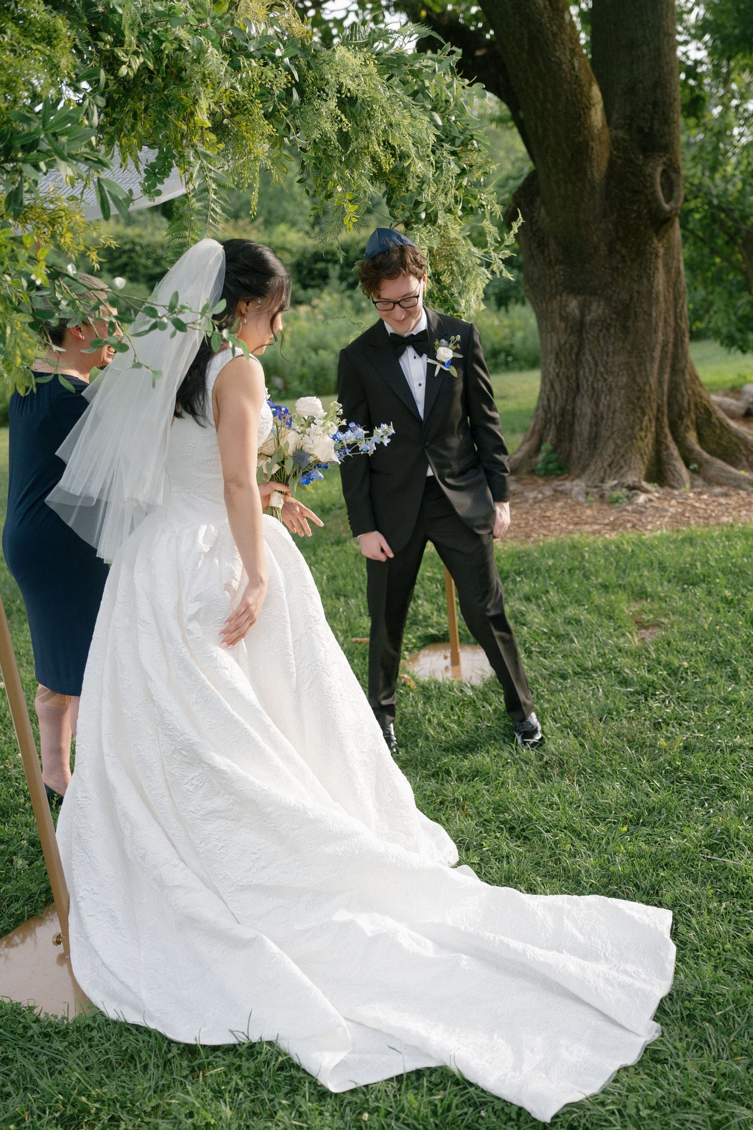 Bride in elegant white gown with long train walks toward smiling groom in black tuxedo during outdoor wedding ceremony.