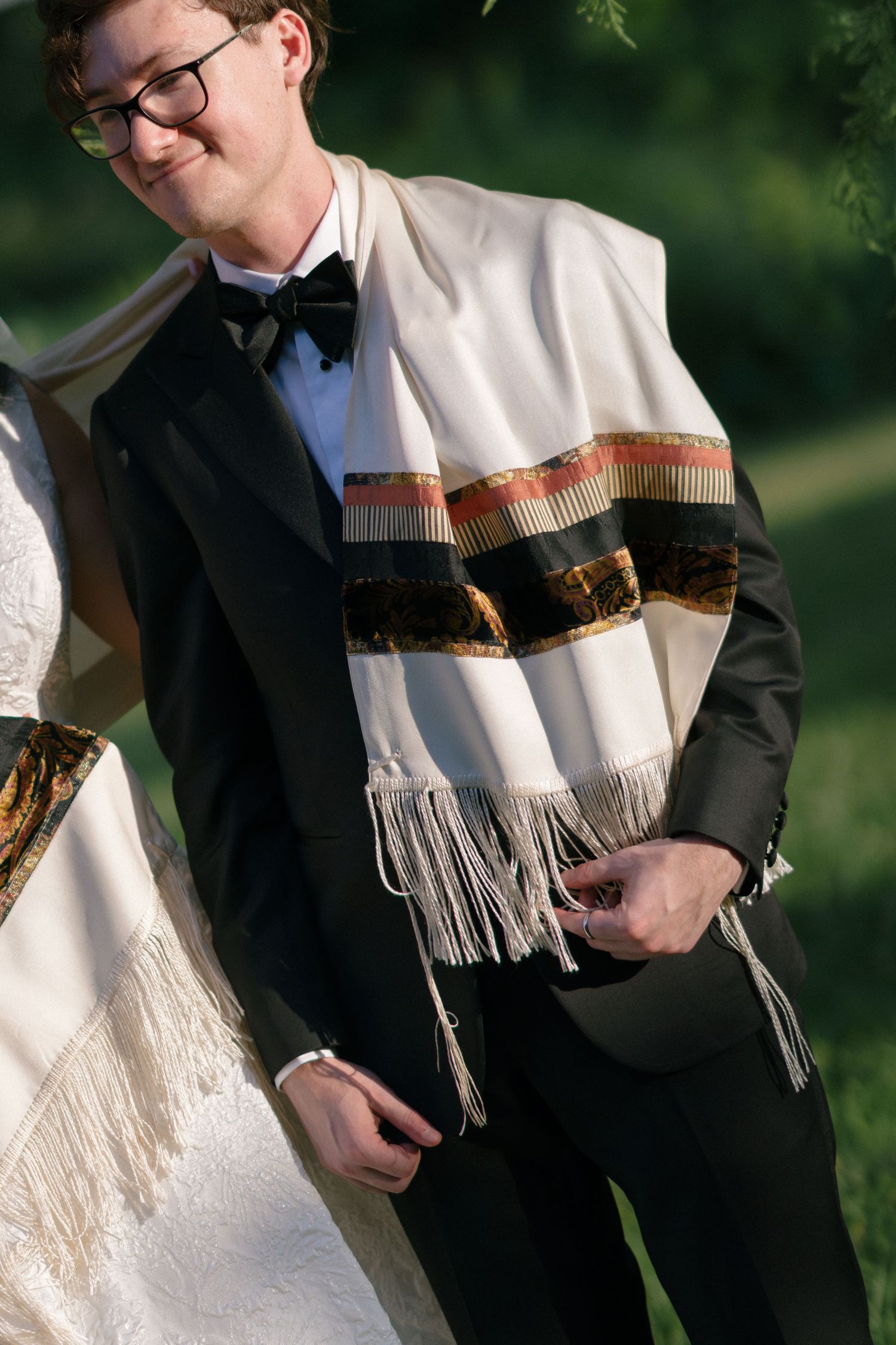 Groom in black tuxedo wearing a traditional Jewish tallit prayer shawl with fringe at outdoor wedding ceremony.