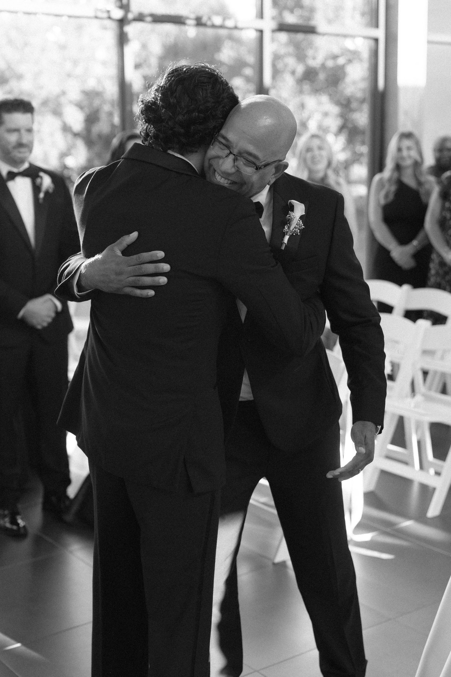 Emotional black and white photo of two men sharing a heartfelt hug during a wedding ceremony.