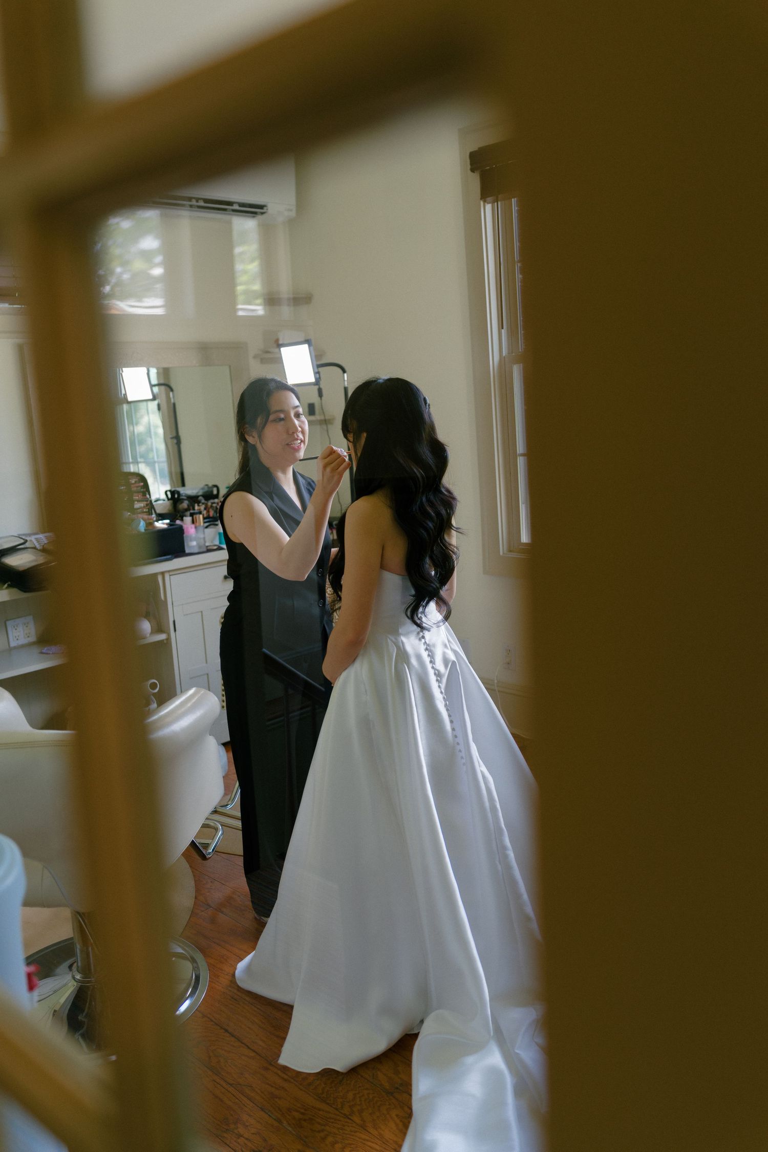Makeup artist applying finishing touches to bride in strapless white ball gown before wedding.