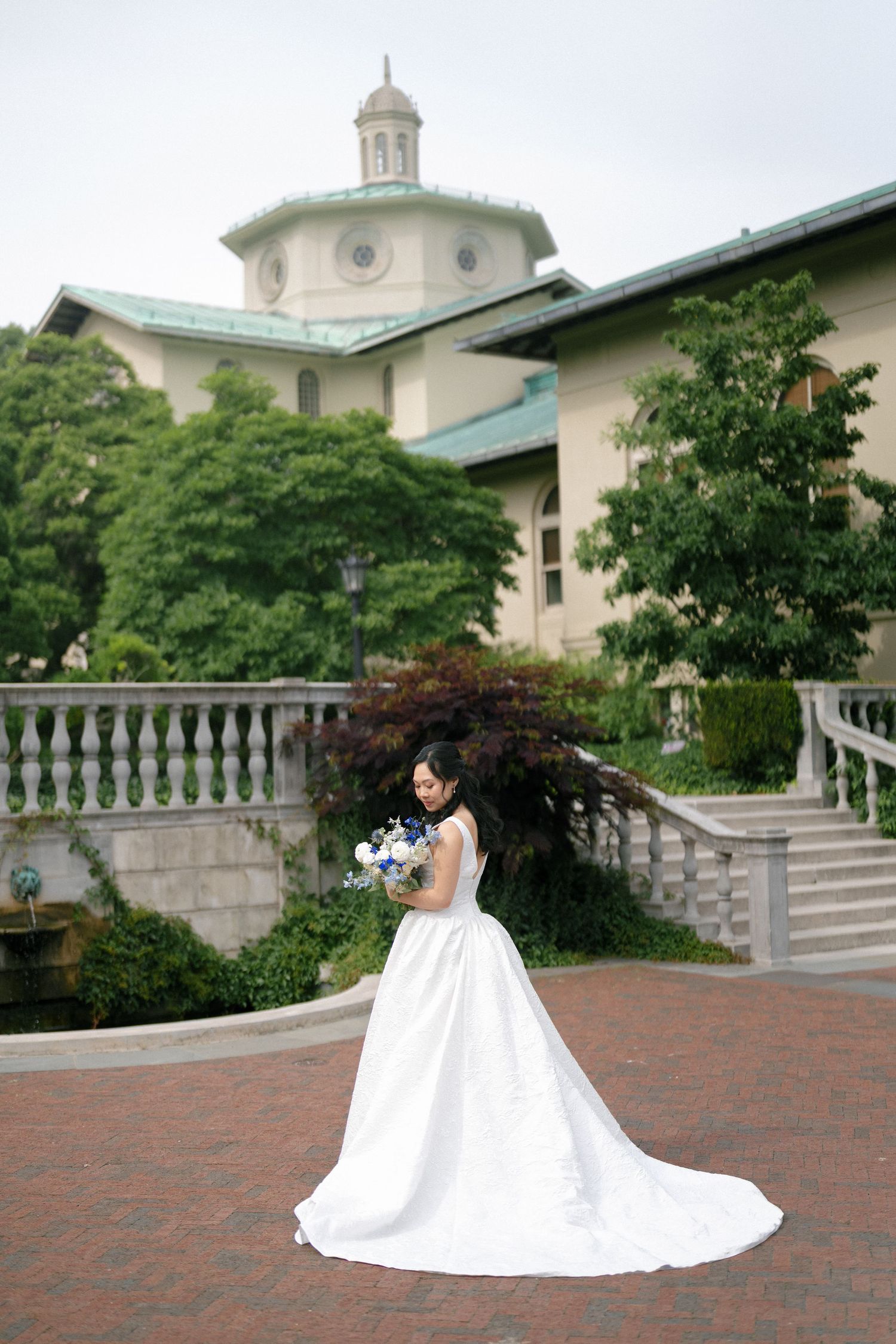 Bride in elegant white ball gown with long train holds bouquet outside grand venue with balustrade.