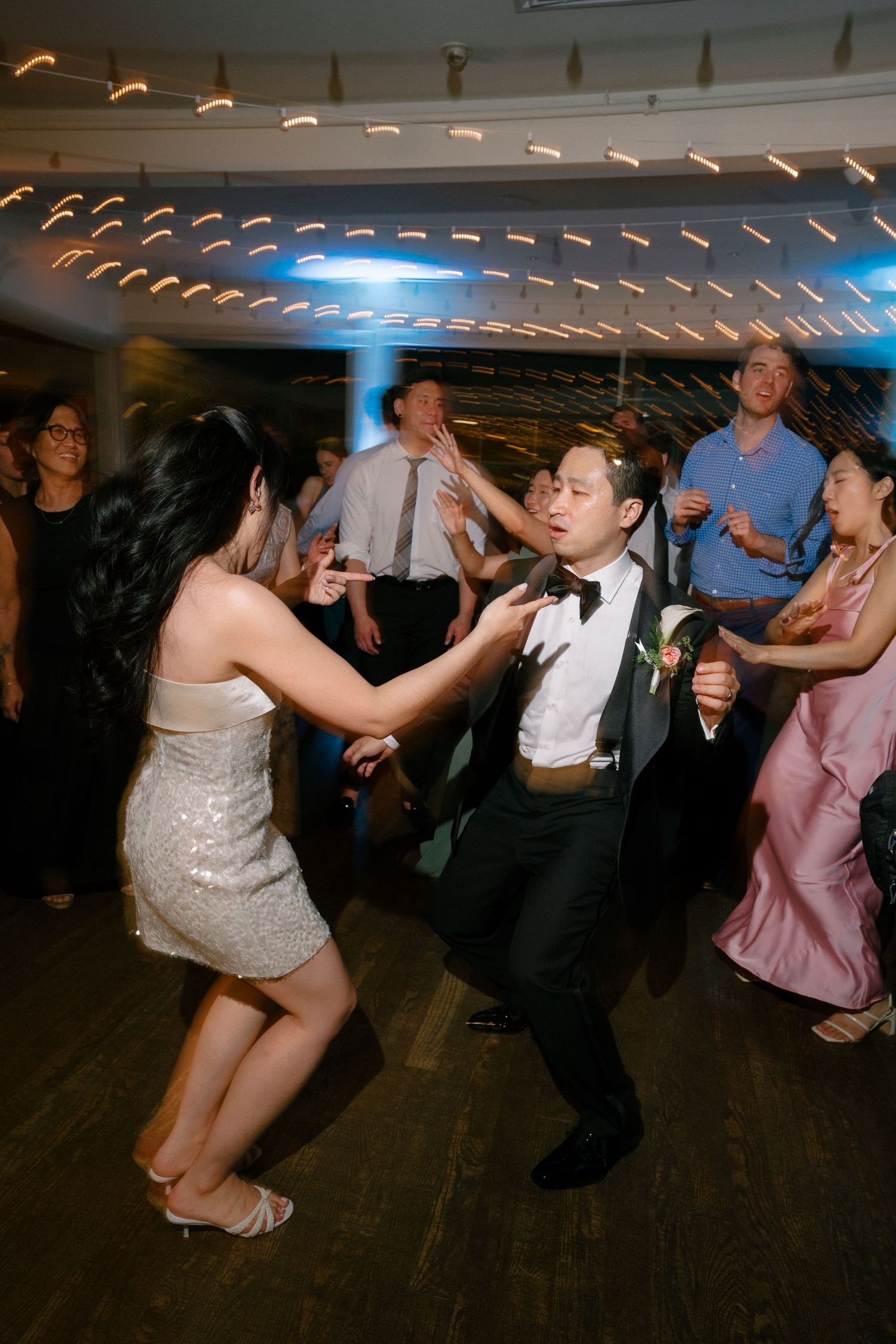 Bride and groom dance energetically on a crowded wedding reception dance floor with string lights above.
