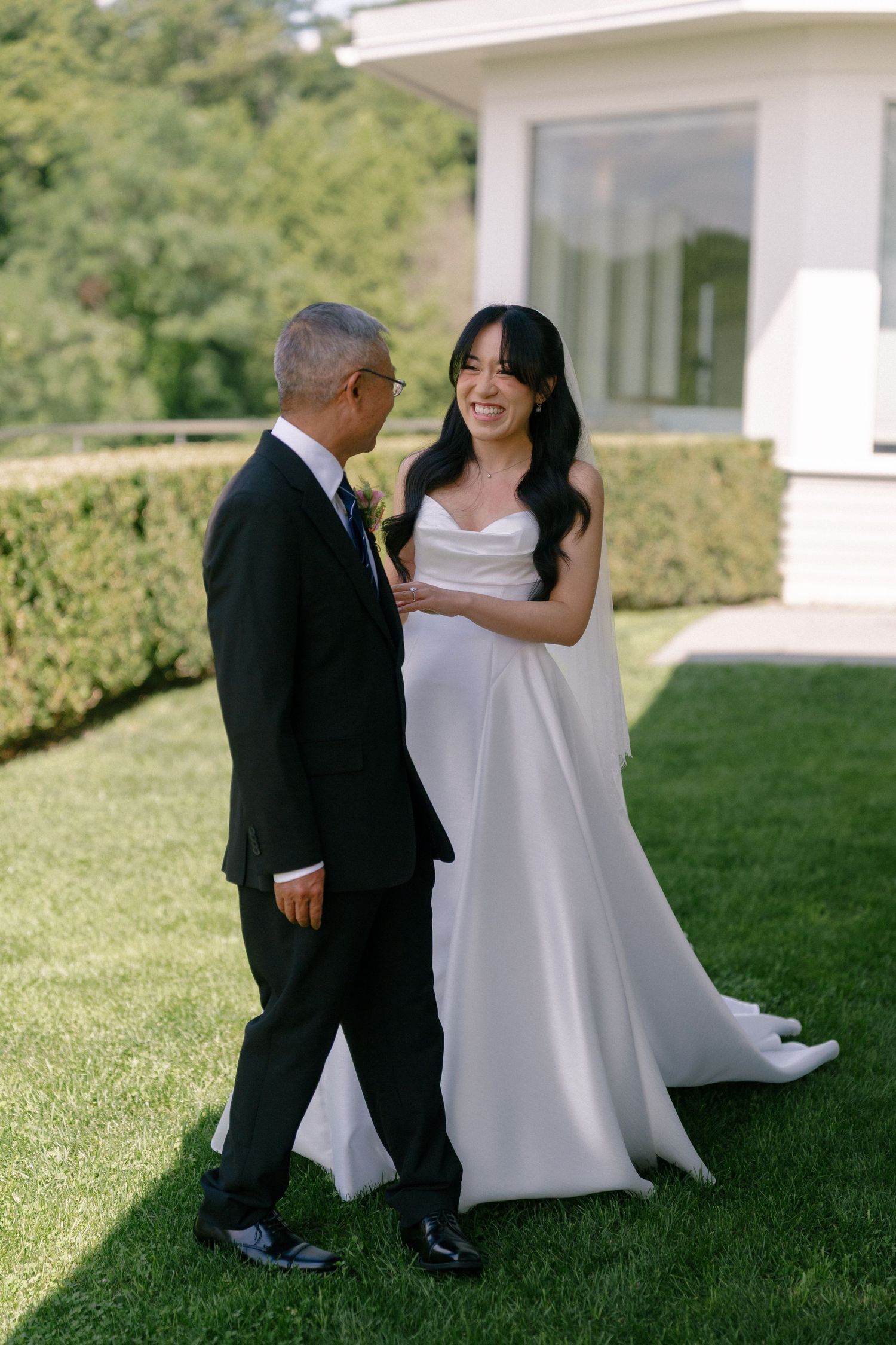 Bride in white cowl neck gown walks with father on green lawn before outdoor wedding ceremony.