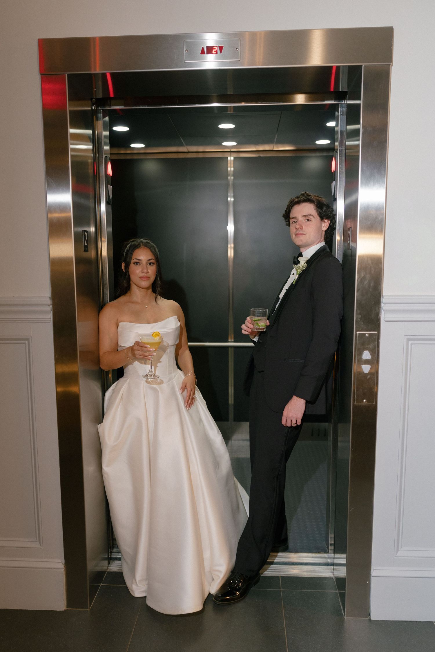 Bride and groom pose with cocktails inside a silver elevator at their wedding venue.