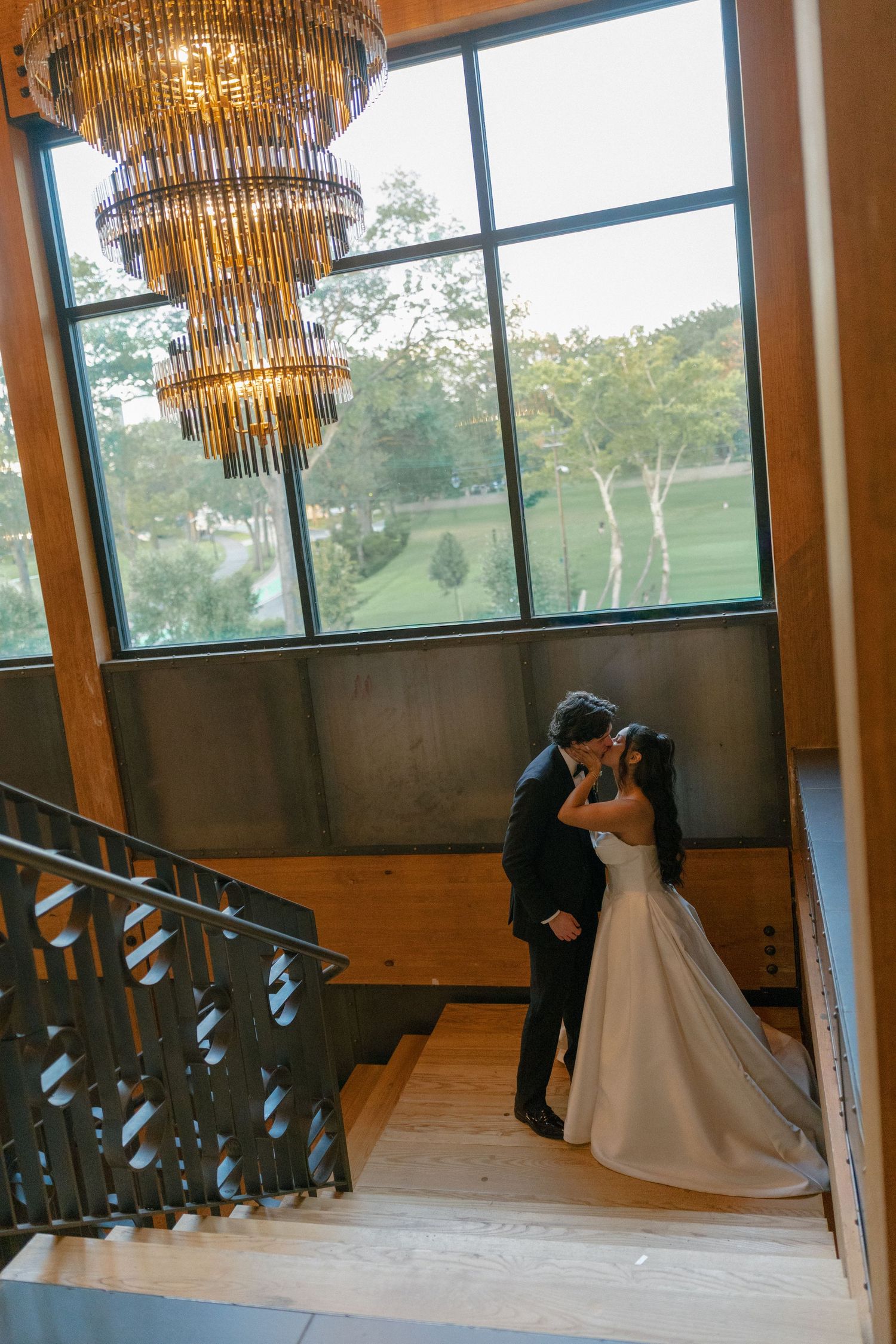 Bride and groom share a kiss on a grand staircase beneath a chandelier with large windows and greenery behind them.